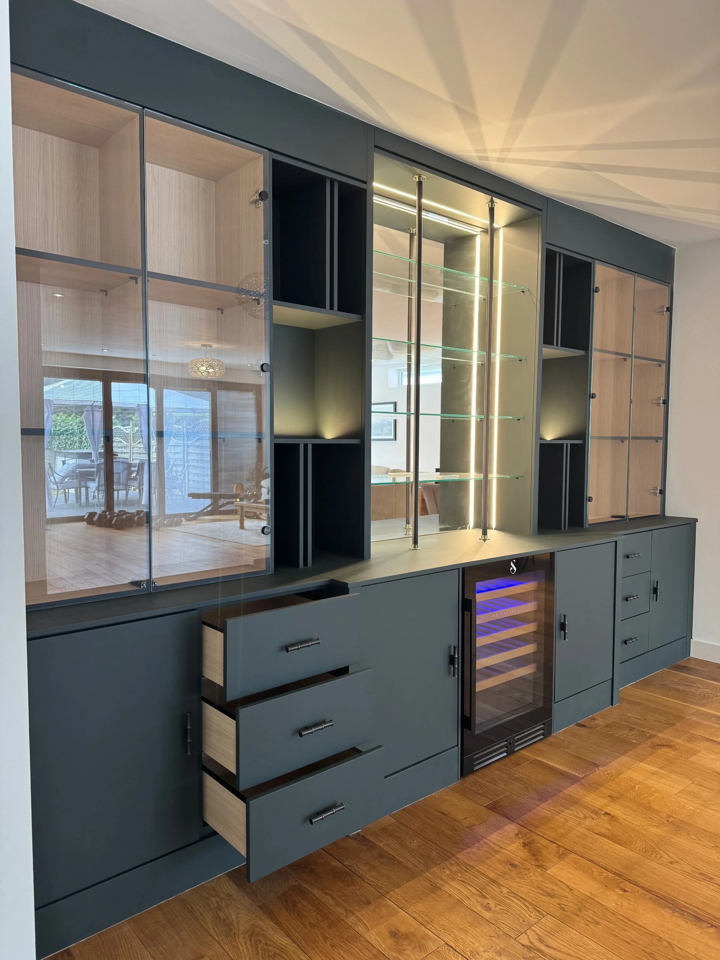 Modern built-in cabinet with glass doors, open shelves, and a wine cooler in a room with wooden flooring.