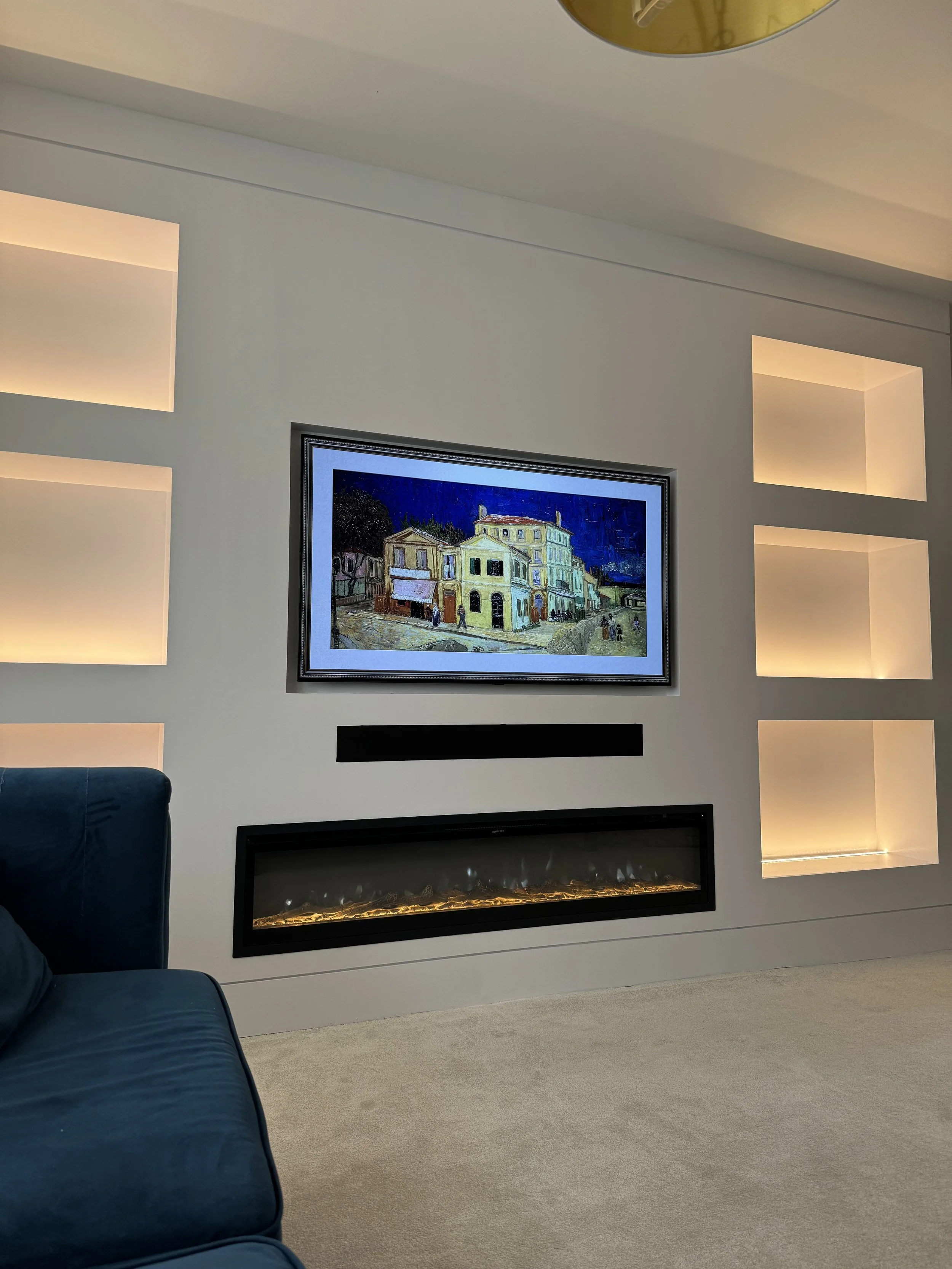 Interior living room with a wall-mounted flat-screen TV displaying artwork of buildings, a modern horizontal fireplace beneath the TV, and illuminated square wall niches on the wall to the right, with a partial view of a blue sofa on the left.
