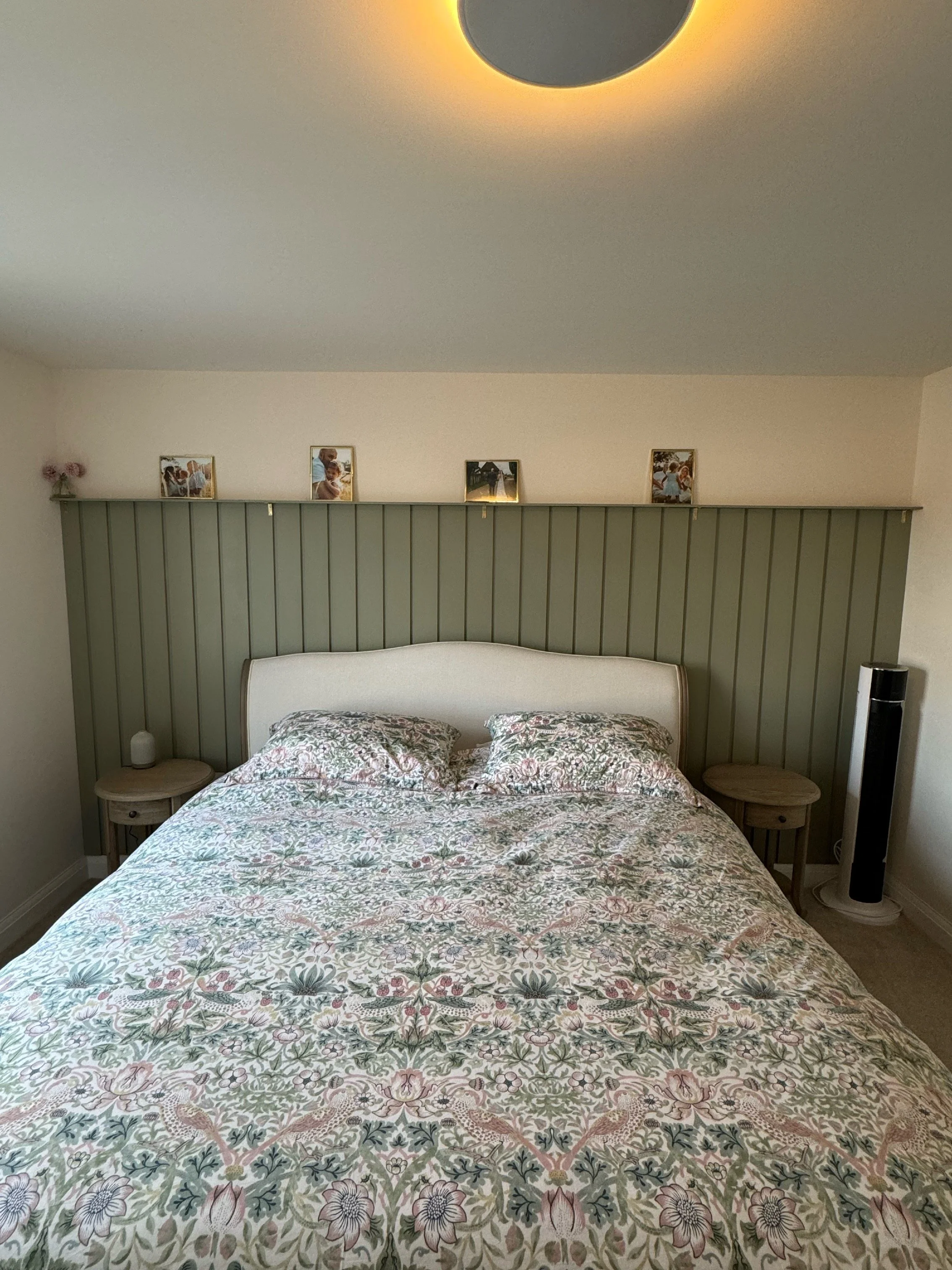A cozy bedroom with a bed, floral bedspread, two matching pillows, a green paneled headboard, two round bedside tables, a small decorative object and a lamp on the left, a tall black fan on the right, and a ceiling light fixture. Photos sit on a shel