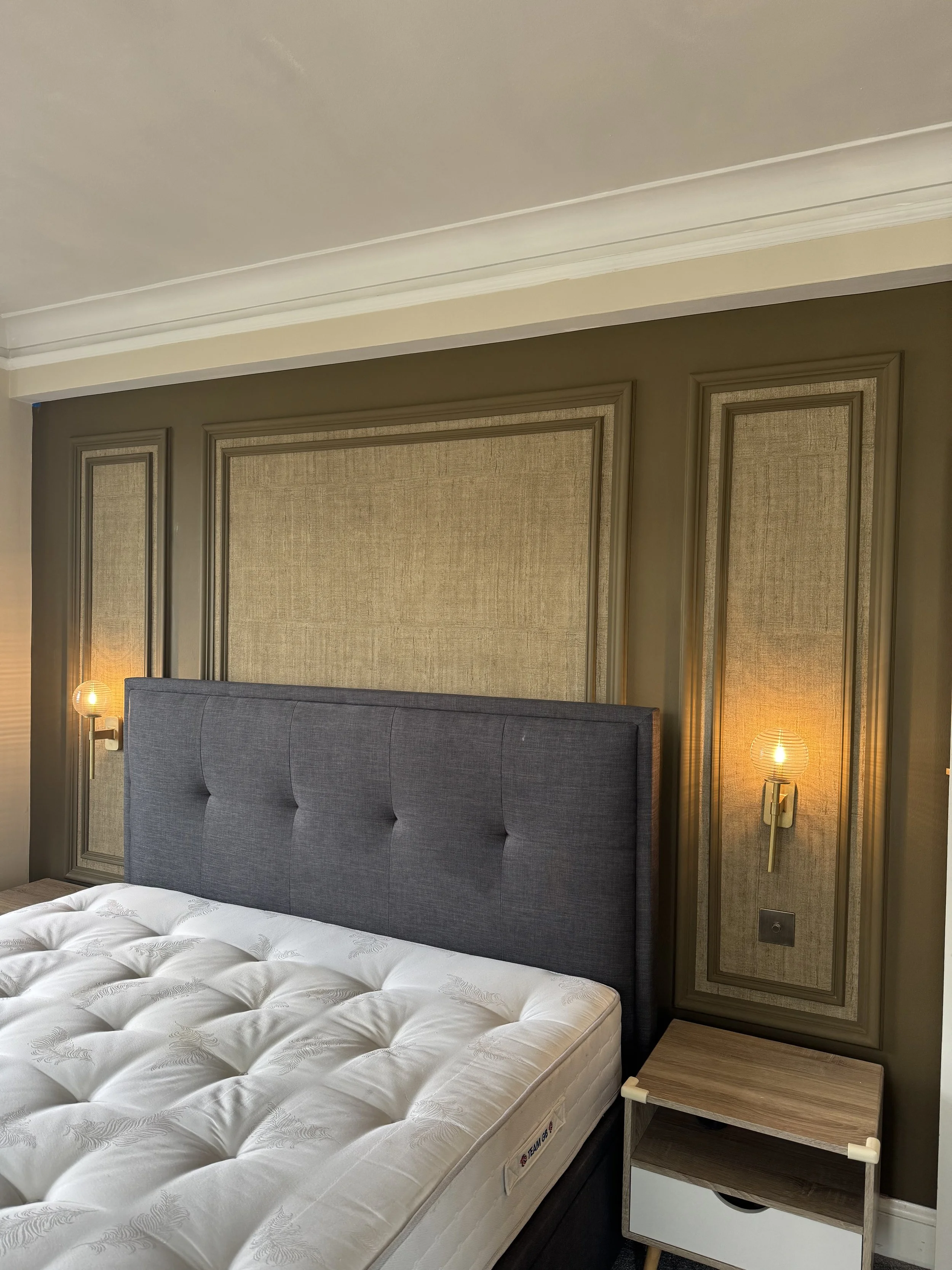 Side view of a bedroom corner with a bed, a cushioned headboard, two wall-mounted lamps, and a small wooden nightstand.