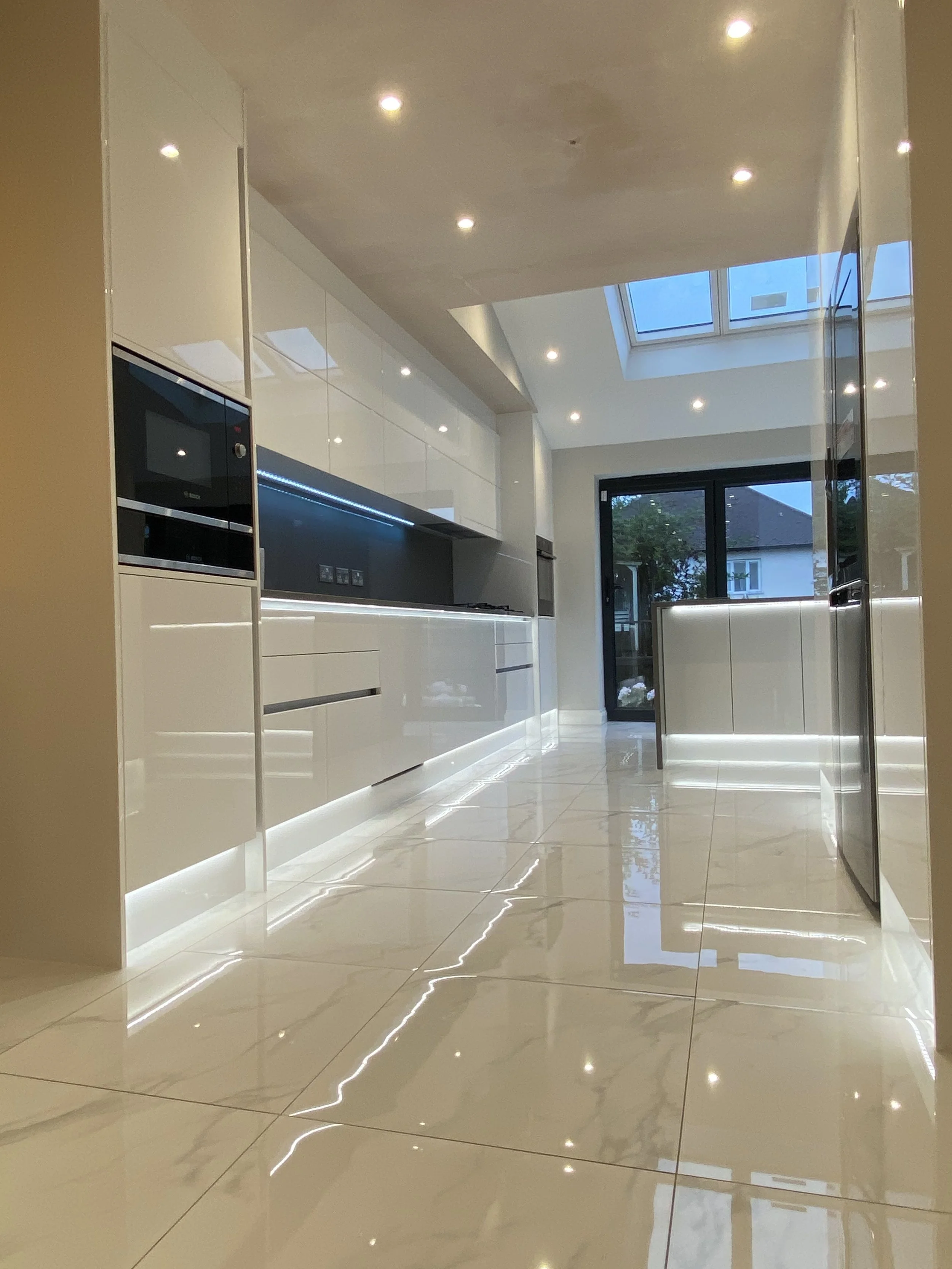 Modern kitchen with white cabinets, black appliances, and a skylight in the ceiling, reflecting light on the marble floor.