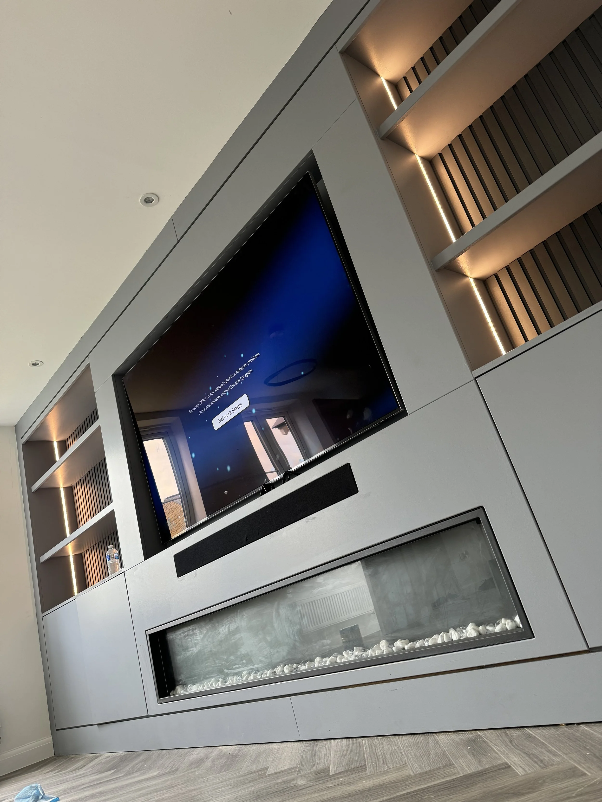 A modern entertainment center with a large flat-screen TV, built-in fireplace with decorative rocks, and shelves with wooden slats and lighting.