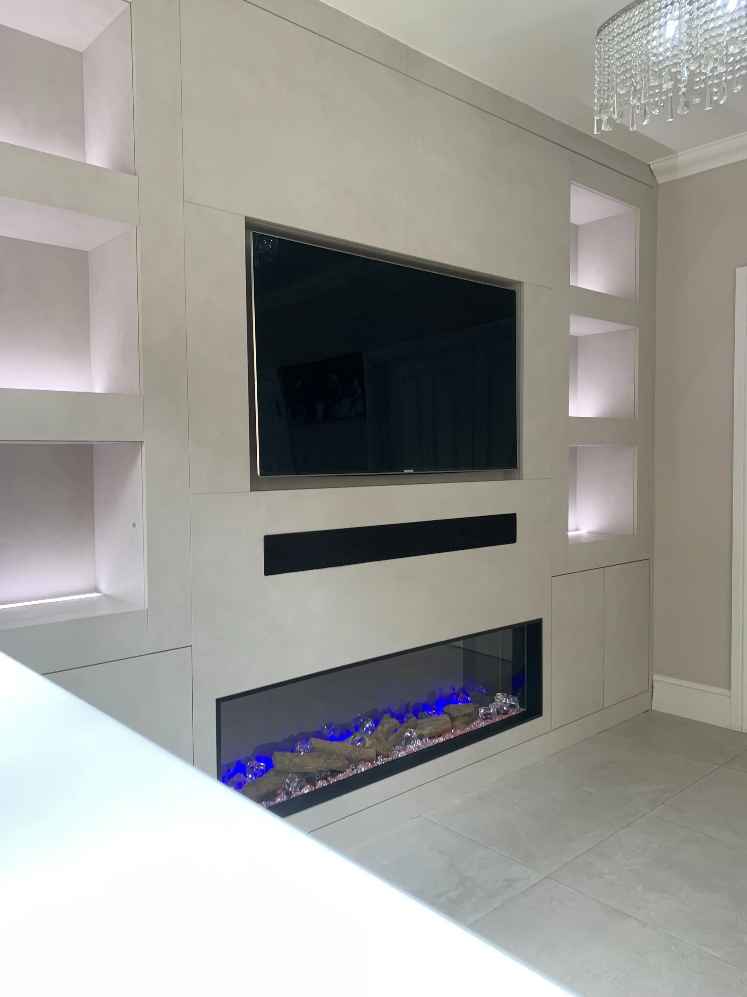 Living room wall with a large flat-screen TV, built-in shelves with lighting, and an electric fireplace with blue flames and decorative stones.