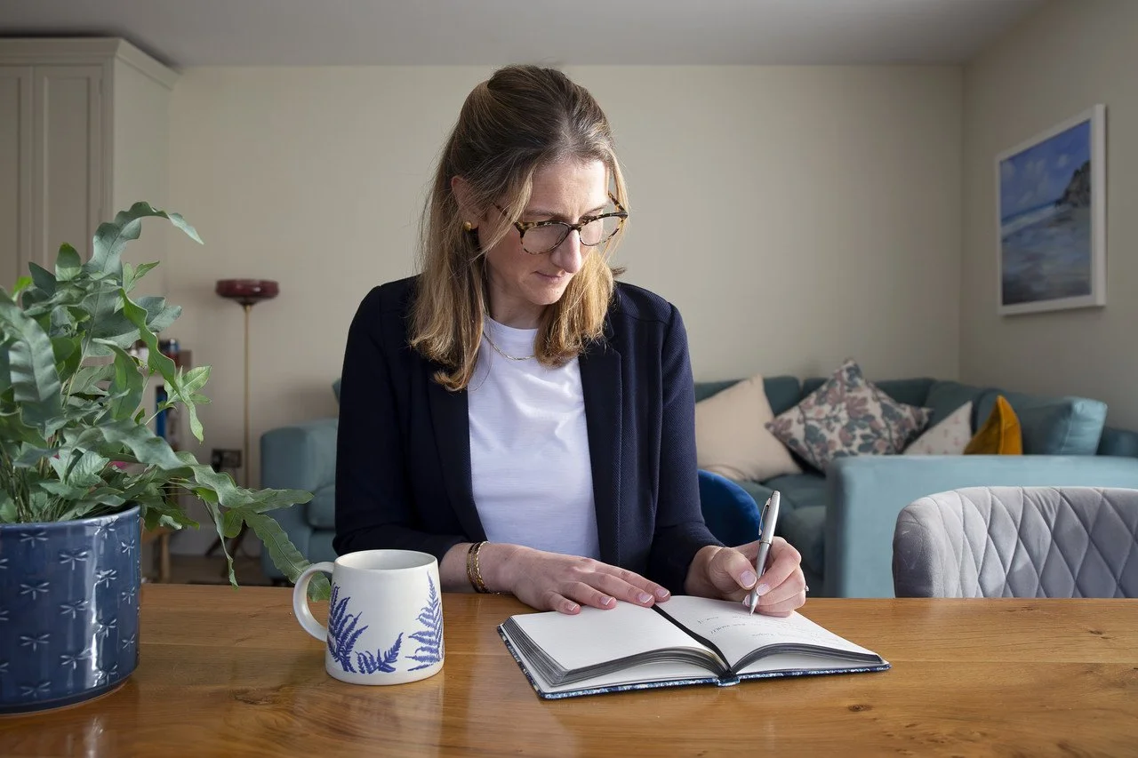Kirsty Murdoch, professional life coach for mid-career professionals, sitting in a relaxed setting making a note of a coaching booking with a cup of tea and plant next to her.