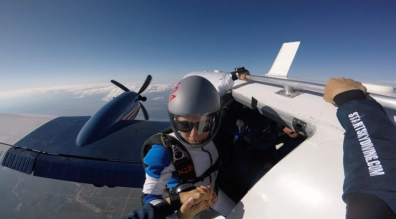 Skydiver exiting an airplane at high altitude with blue sky and clouds below.