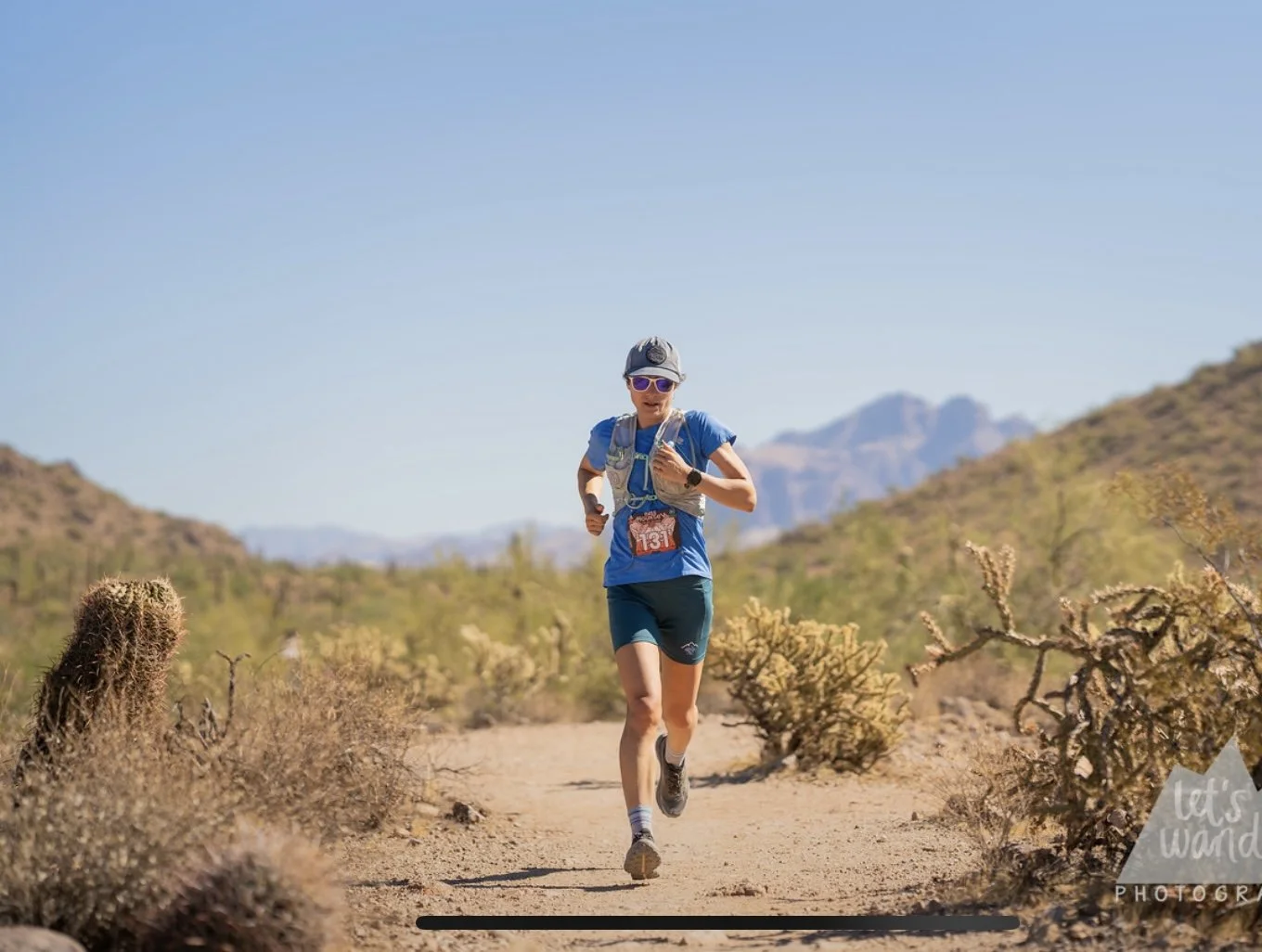 A woman running fast on a desert trail surrounded by cacti and hills under a clear blue sky.