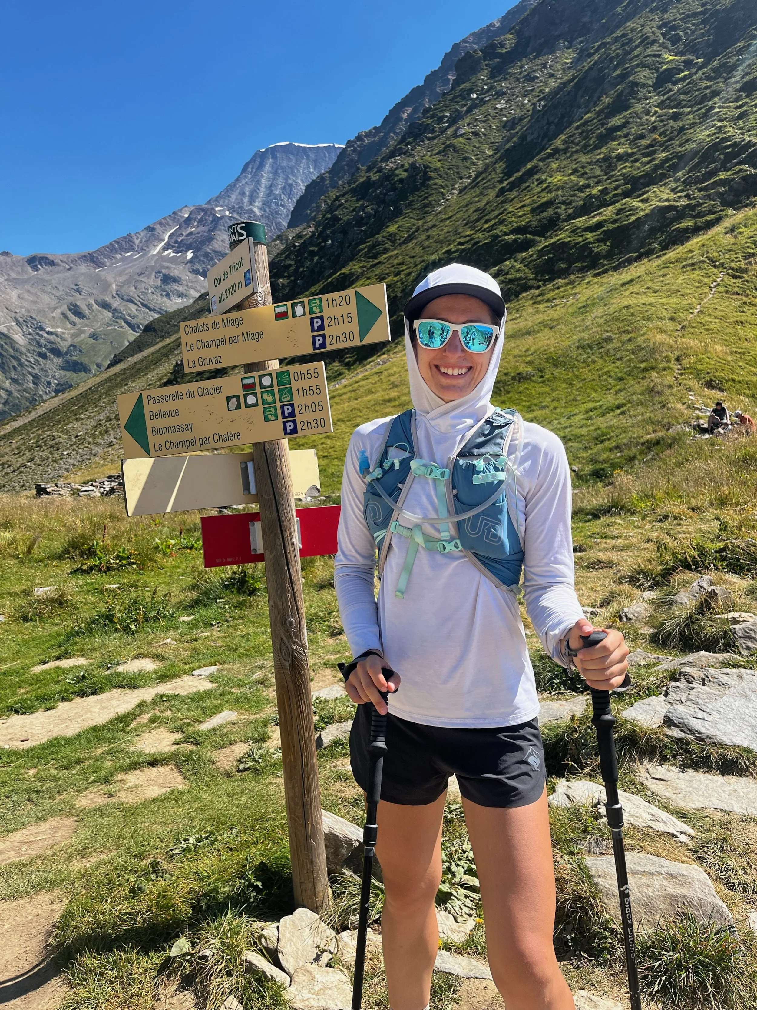 A smiling woman in outdoor hiking gear with sunglasses and trekking poles standing next to a trail sign in a mountainous area along the trail du mount blanc on a sunny day.