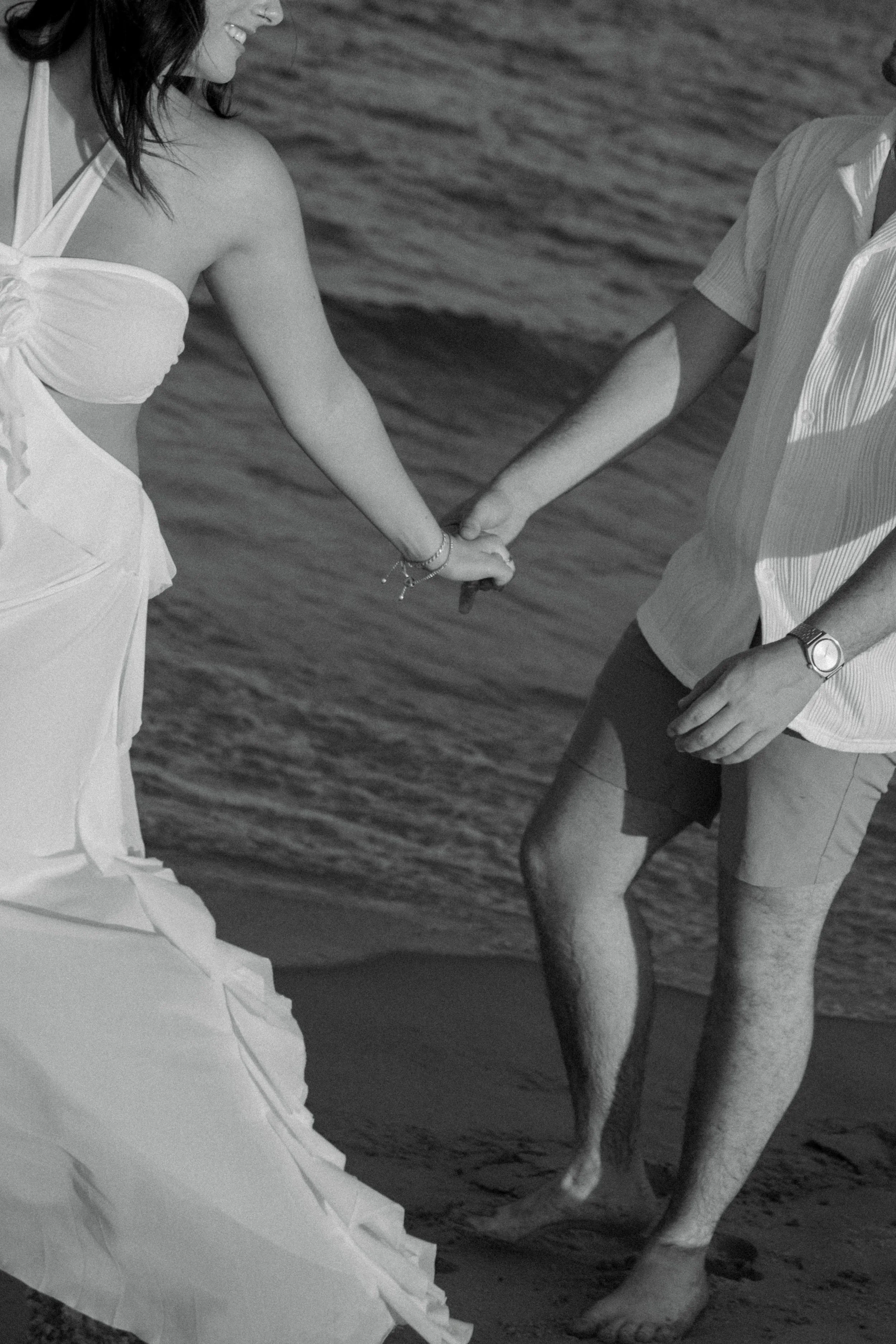 A couple holding hands on the beach, with the ocean in the background. The woman is wearing a flowing dress and the man is dressed casually in a striped shirt and shorts.