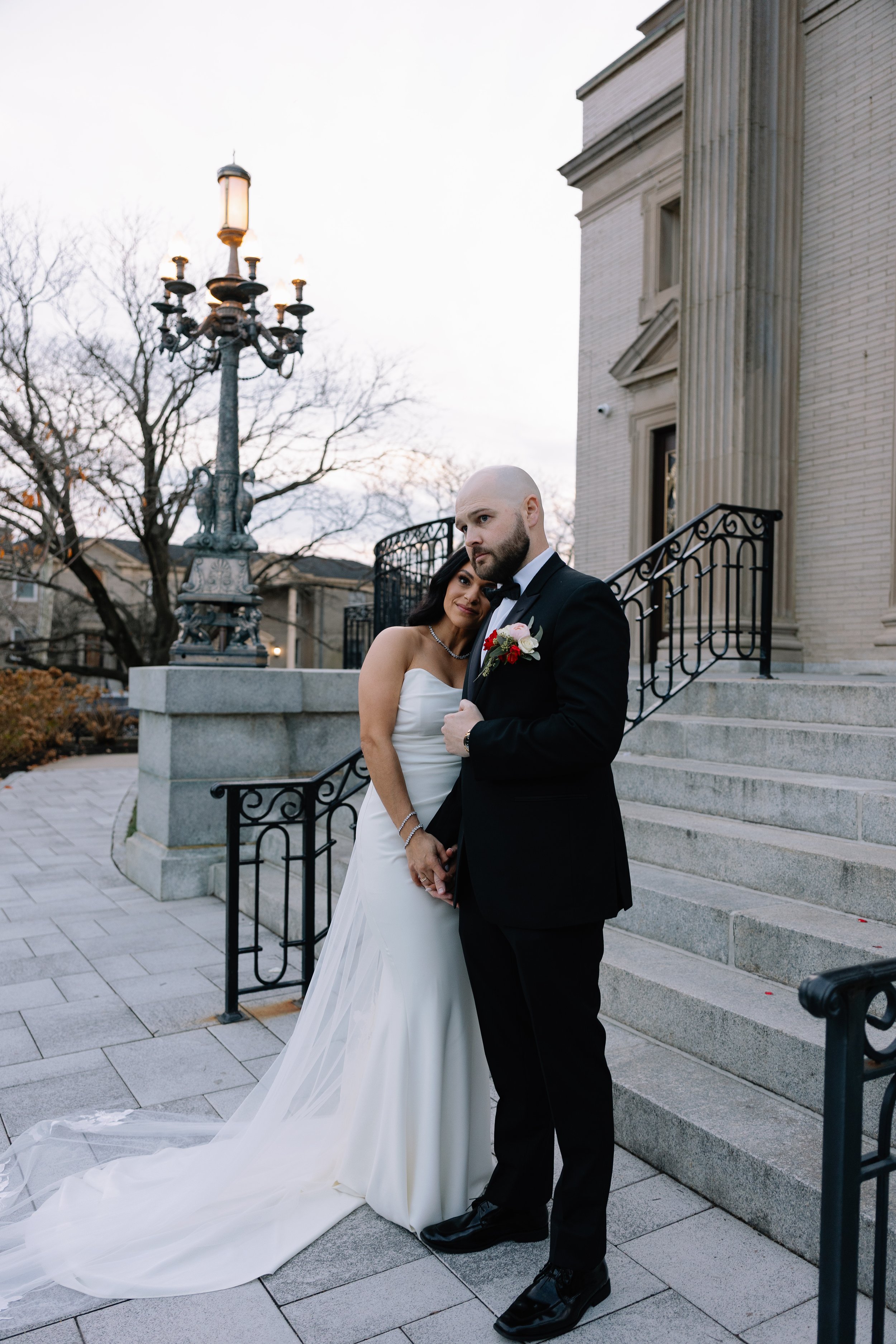 A newlywed couple standing outside a historic building, the bride in a strapless white wedding gown with a train, and the groom in a black tuxedo with a boutonniere, during dusk.