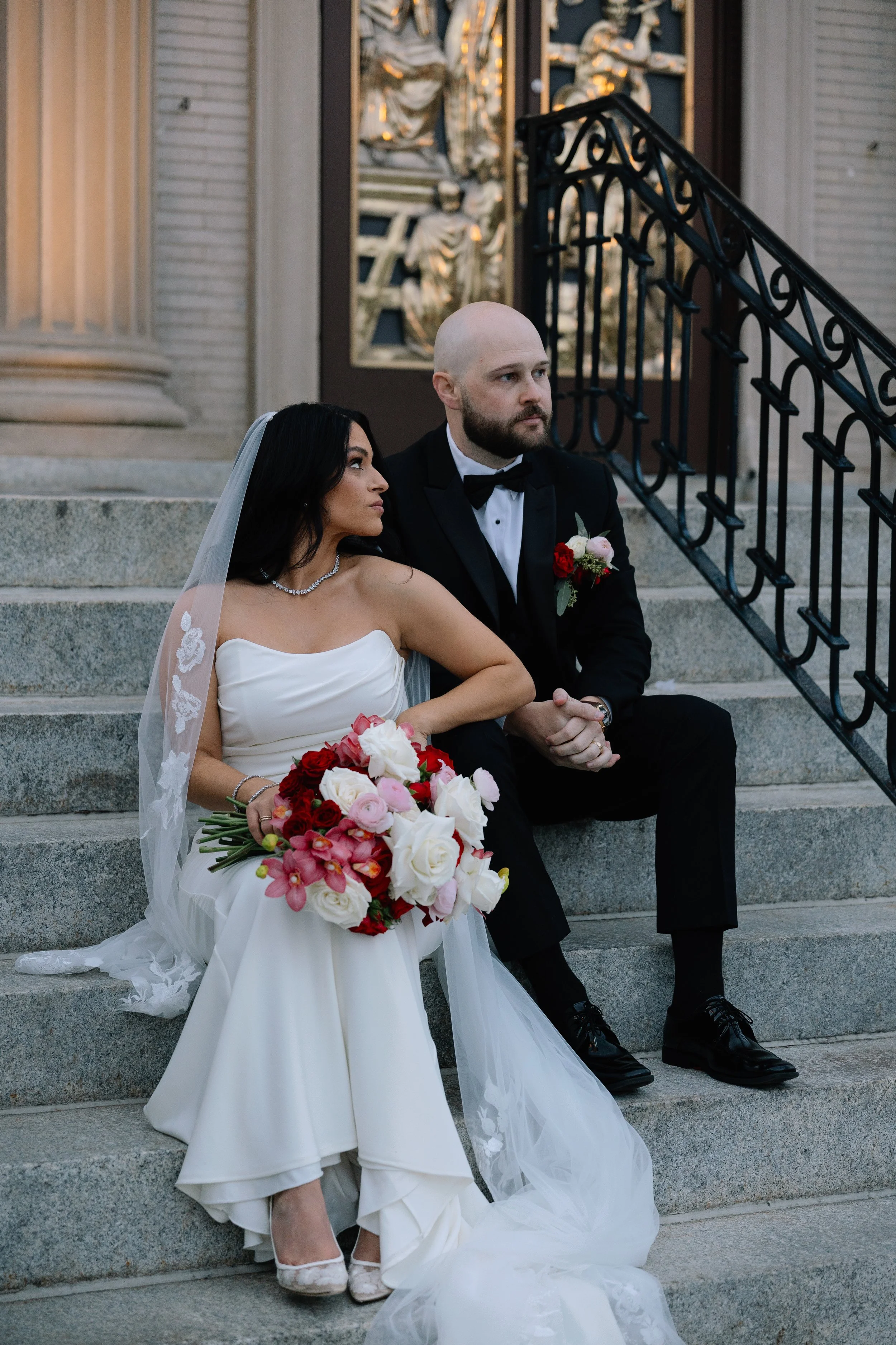 Bride and groom sitting on steps outside a building with ornate window decorations, the bride holding a large bouquet of red, white, and pink flowers, both dressed in formal wedding attire.