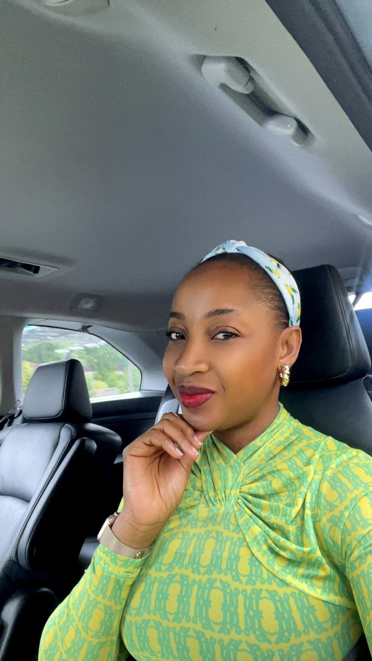 A woman sitting in a car, wearing a yellow and green dress with a white headband and gold earrings, with her hand resting on her chin and a slight smile.
