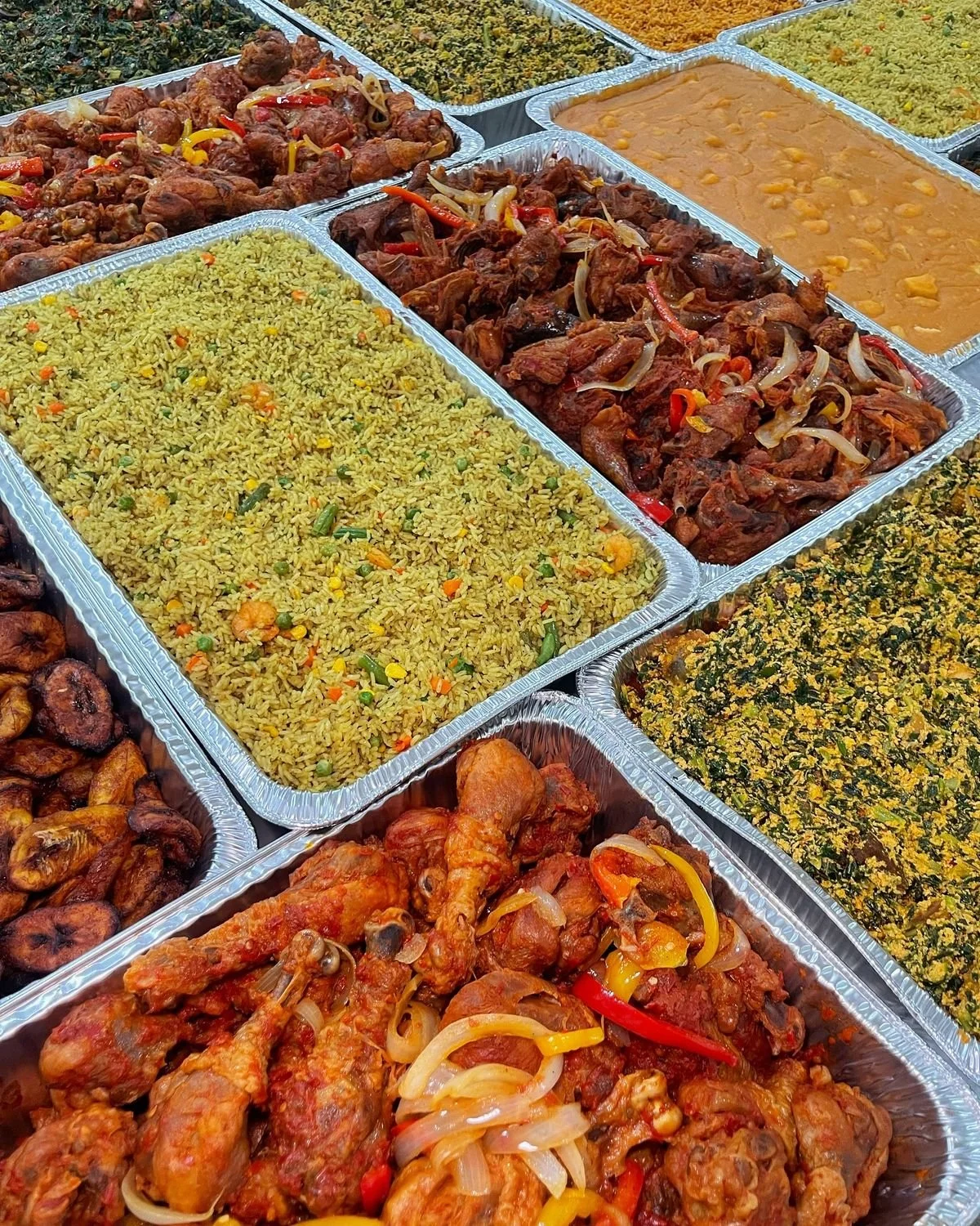 A variety of Indian dishes in aluminum trays, including yellow rice with vegetables, chicken curry, beef curry with onions and peppers, a spinach dish, and other cooked vegetables.