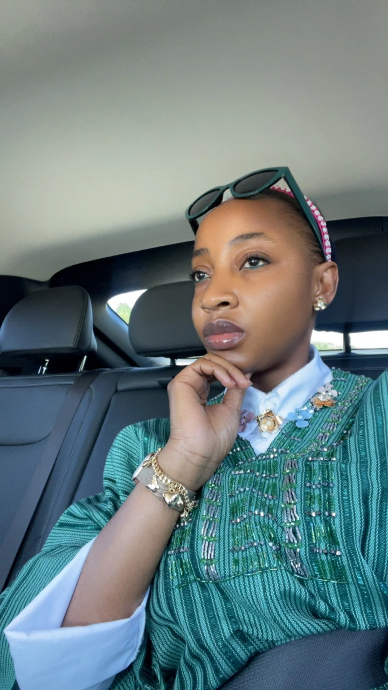 A woman sitting in the backseat of a car, wearing sunglasses on her head, a green patterned jacket, and jewelry, gazing thoughtfully out the window.