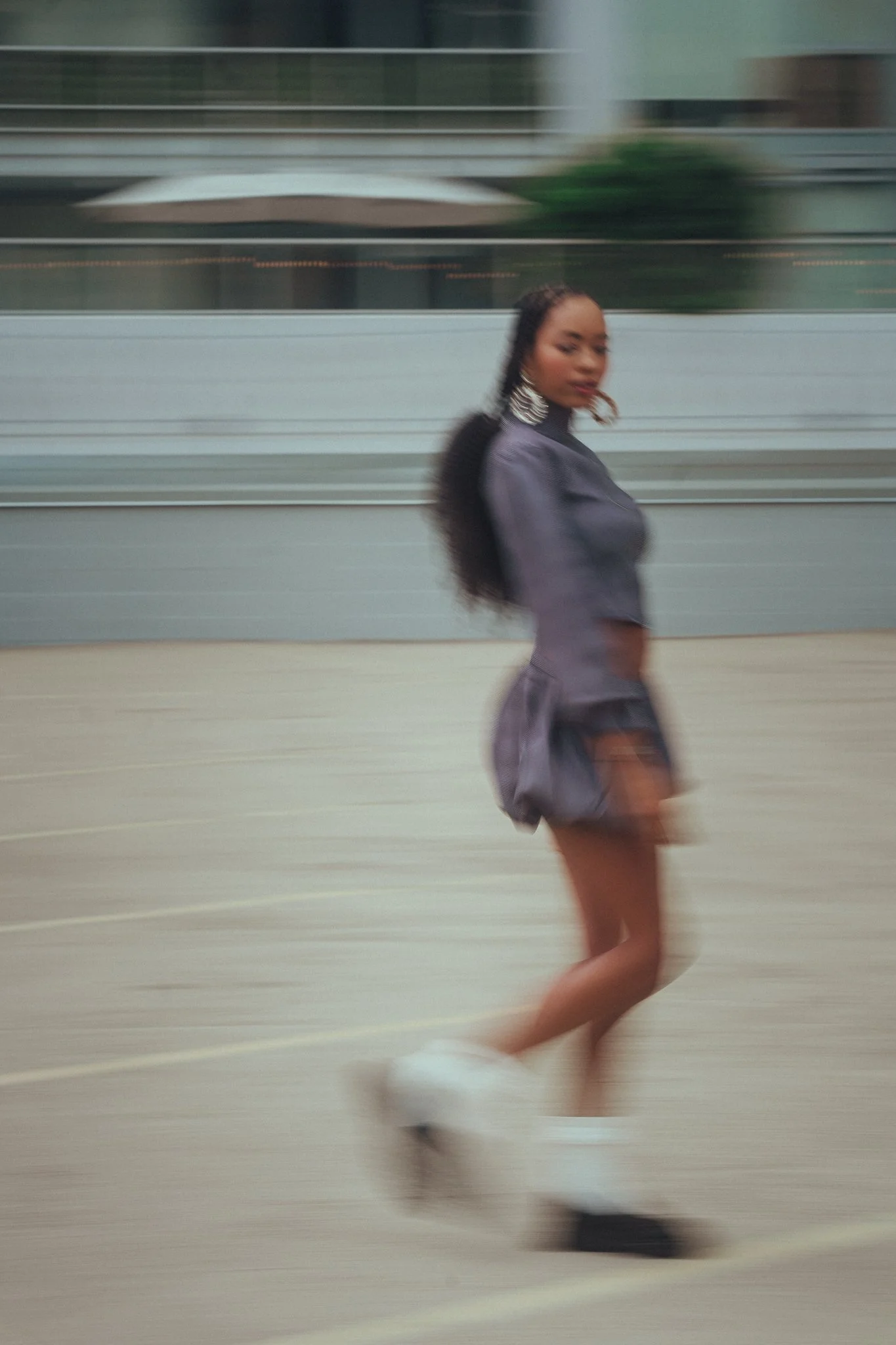 A woman wearing a gray outfit and large earrings is walking on an outdoor paved surface, with motion blur indicating movement.