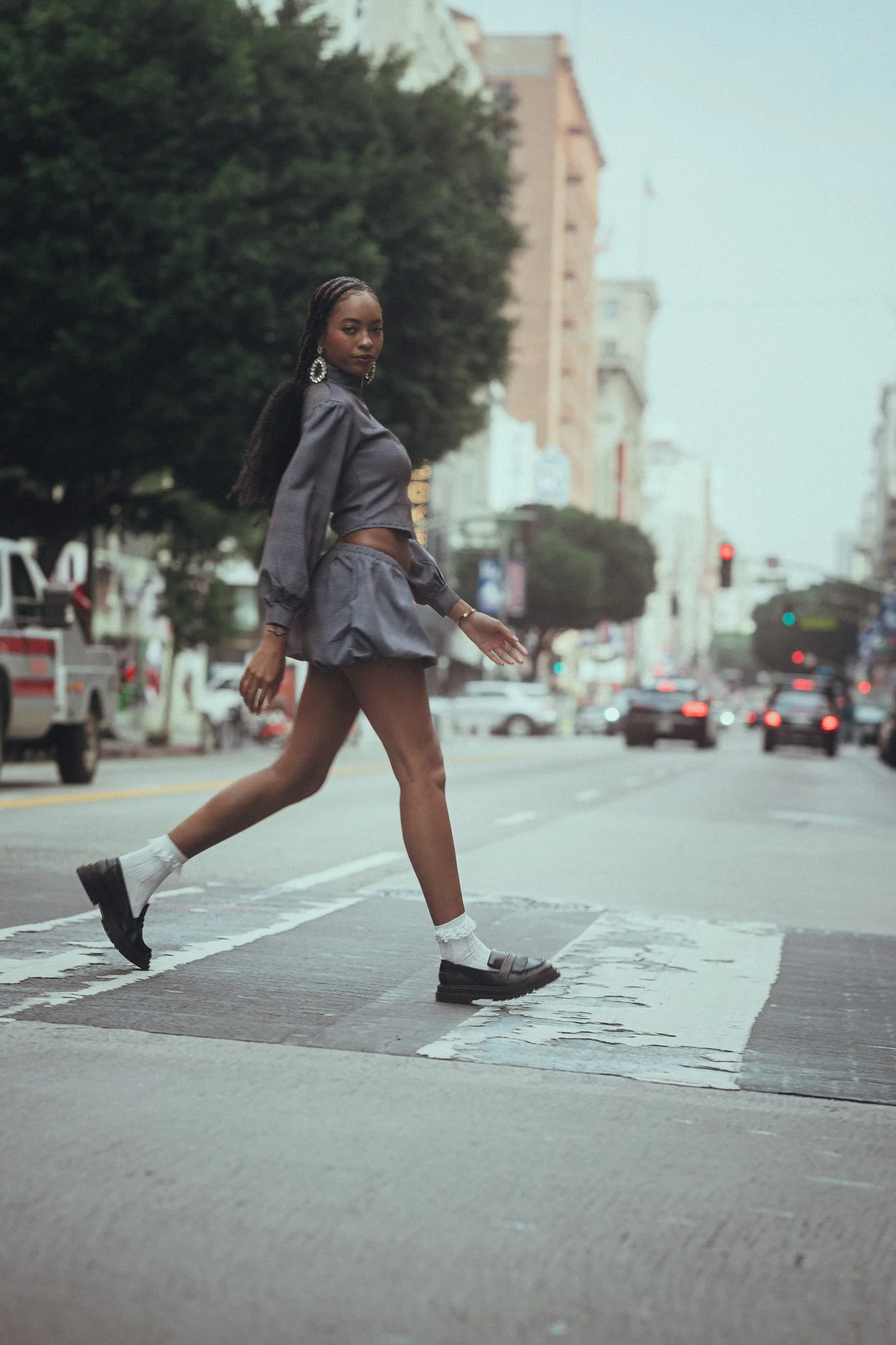 A young woman crossing a city street at a crosswalk, wearing a matching gray satin-like two-piece outfit with puffed sleeves and a ruffled skirt, black shoes, white socks, and large hoop earrings, with trees and buildings in the background.