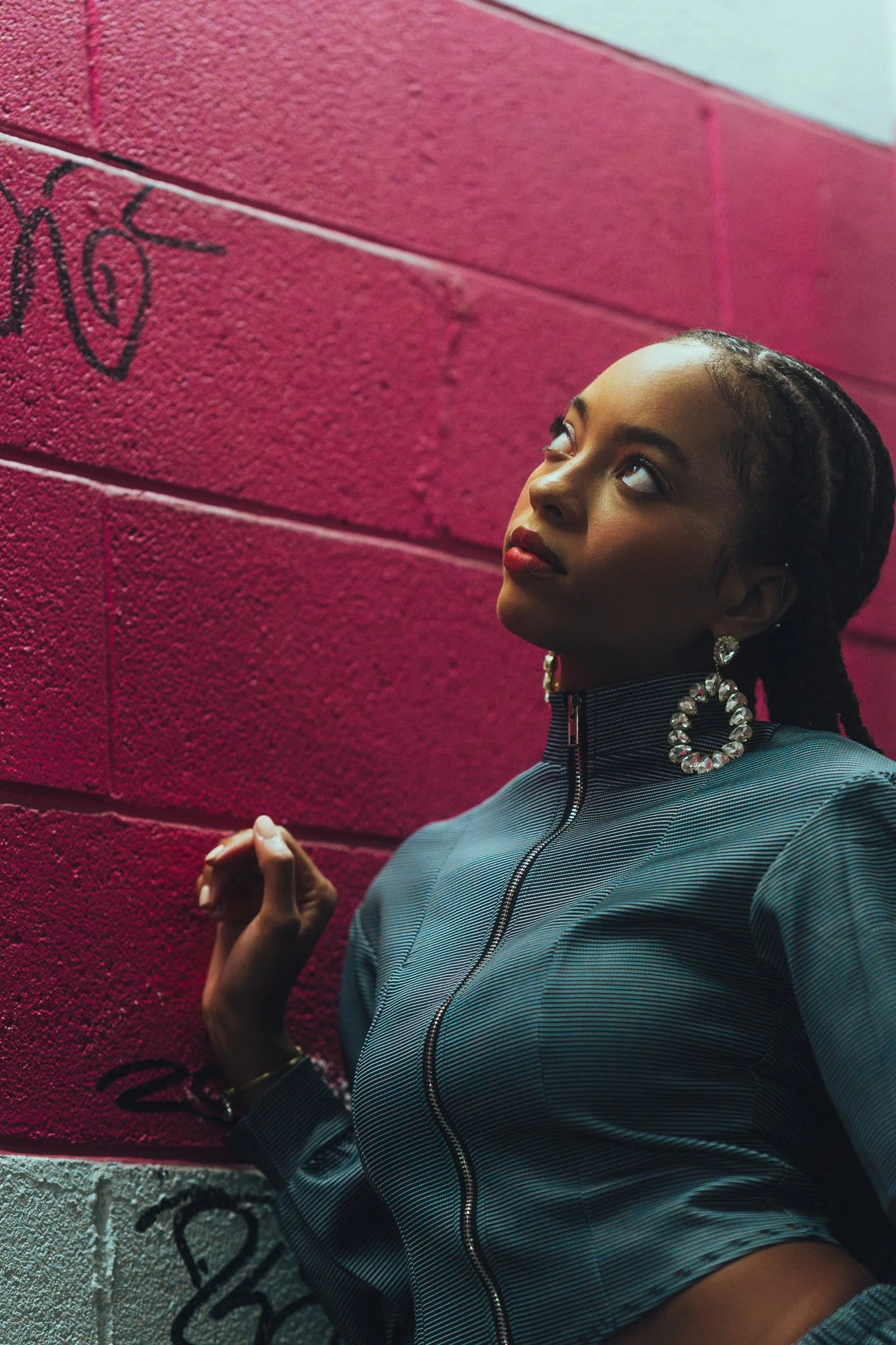 A woman with braided hair, wearing large decorative earrings and a striped zip-up jacket, standing against a red brick wall with graffiti.