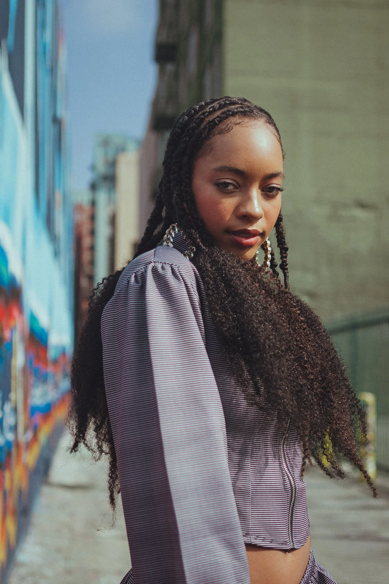 Portrait of a woman with braided hair and hoop earrings, standing in front of a colorful mural on an urban street.