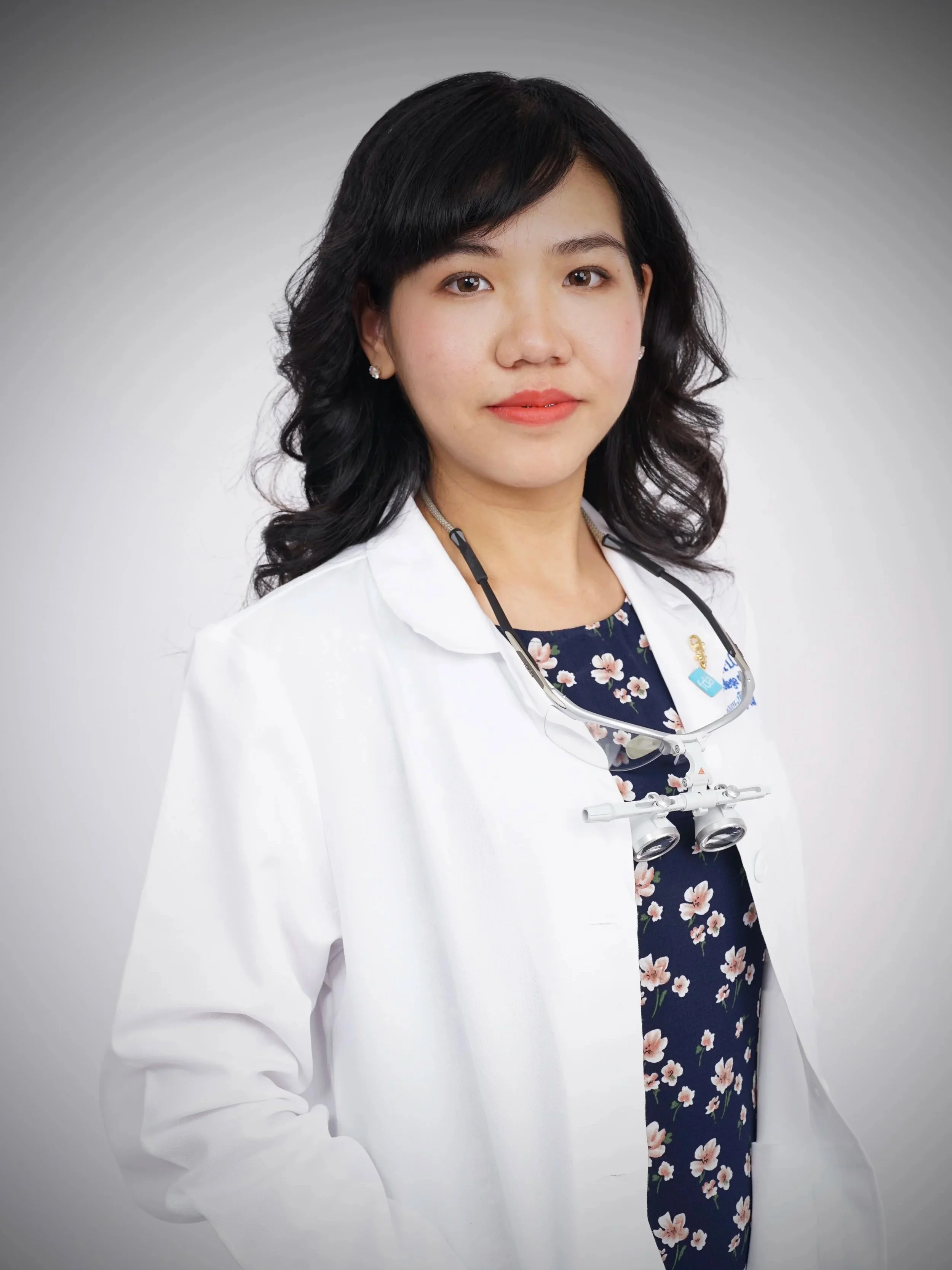 A woman with dark, curled hair wearing a white lab coat and a floral blouse, with a stethoscope around her neck, against a neutral background.