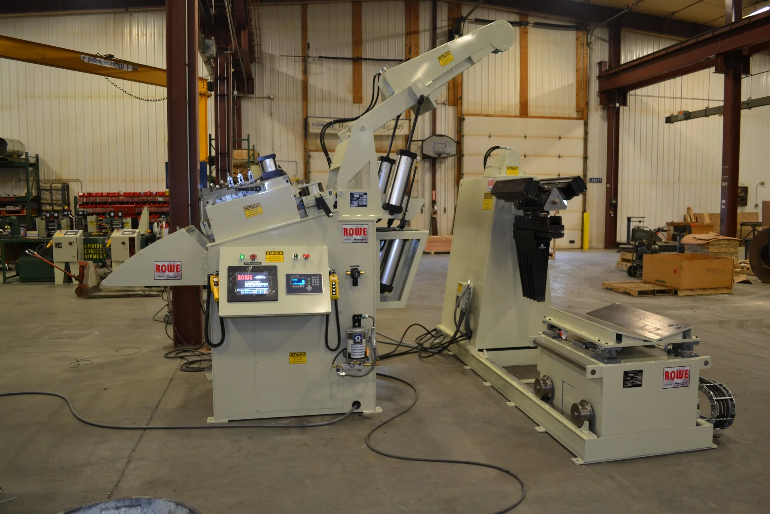 Industrial robotic welding machine labeled 'ROWE' inside a warehouse with various industrial equipment and supplies in the background.