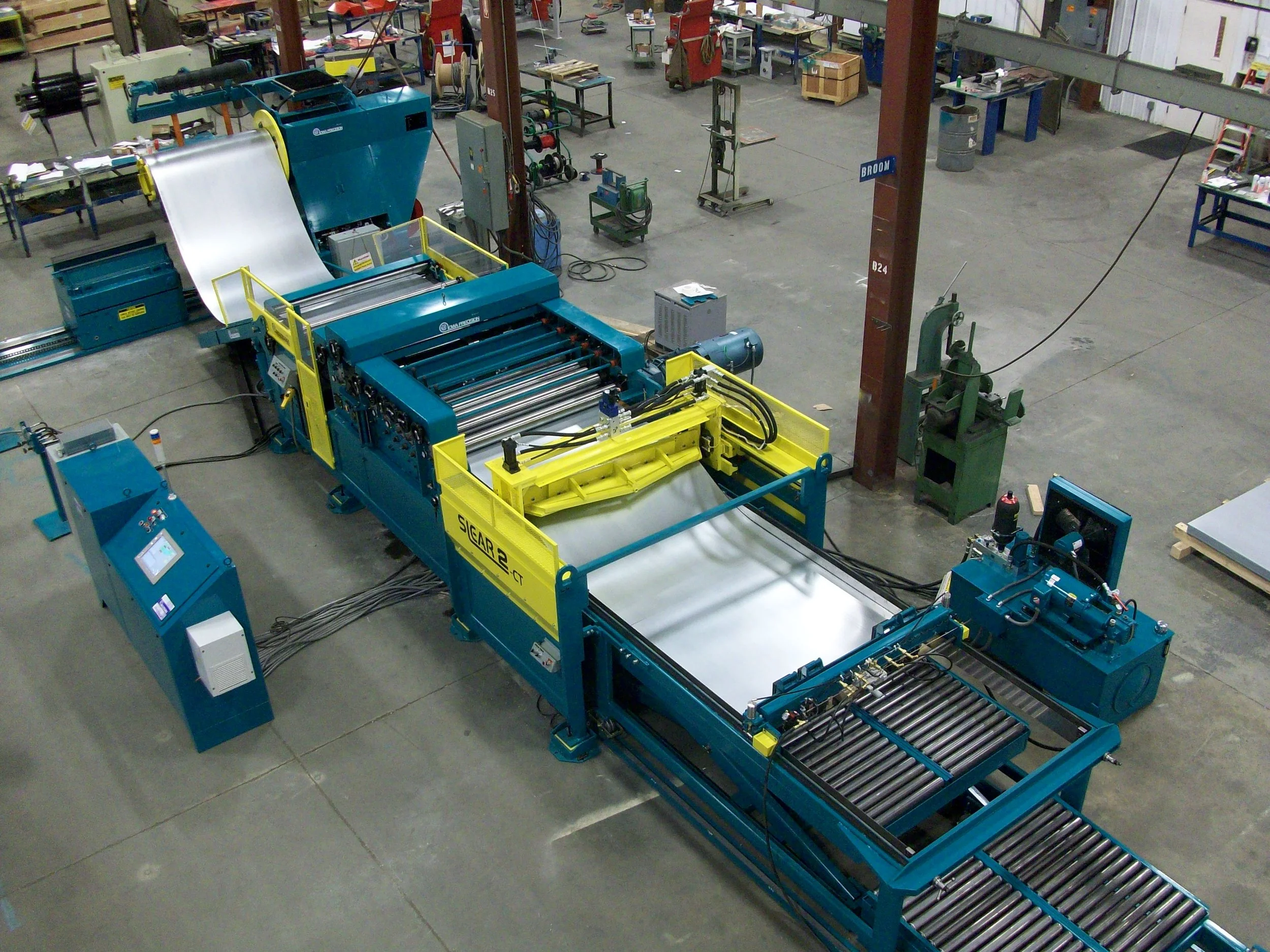 Industrial manufacturing facility with large blue and yellow machinery, rollers, control panels, and storage shelves in the background.