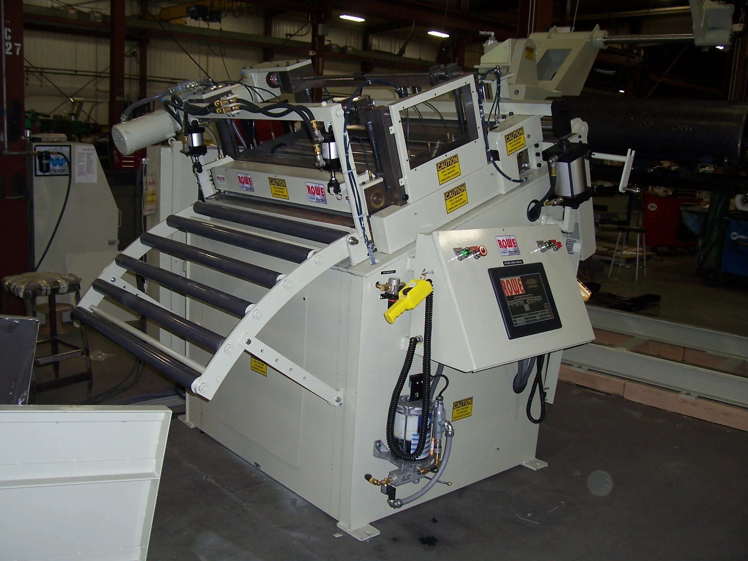 A large industrial machine with rollers, hydraulic components, and a touchscreen control panel in a warehouse or manufacturing setting.