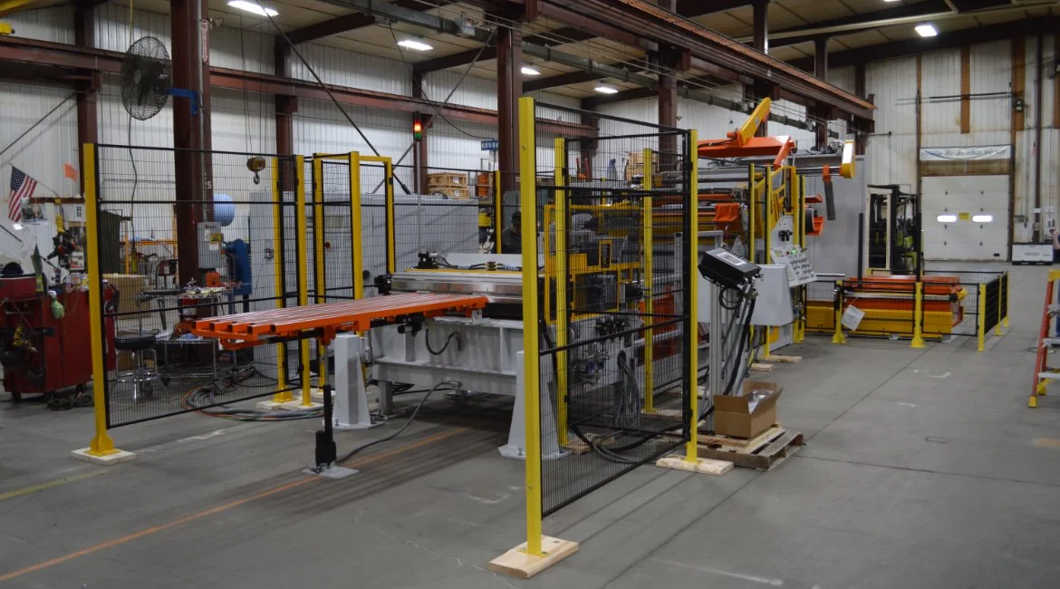 Industrial manufacturing equipment in a factory, surrounded by safety barriers, with various machinery and tools visible.