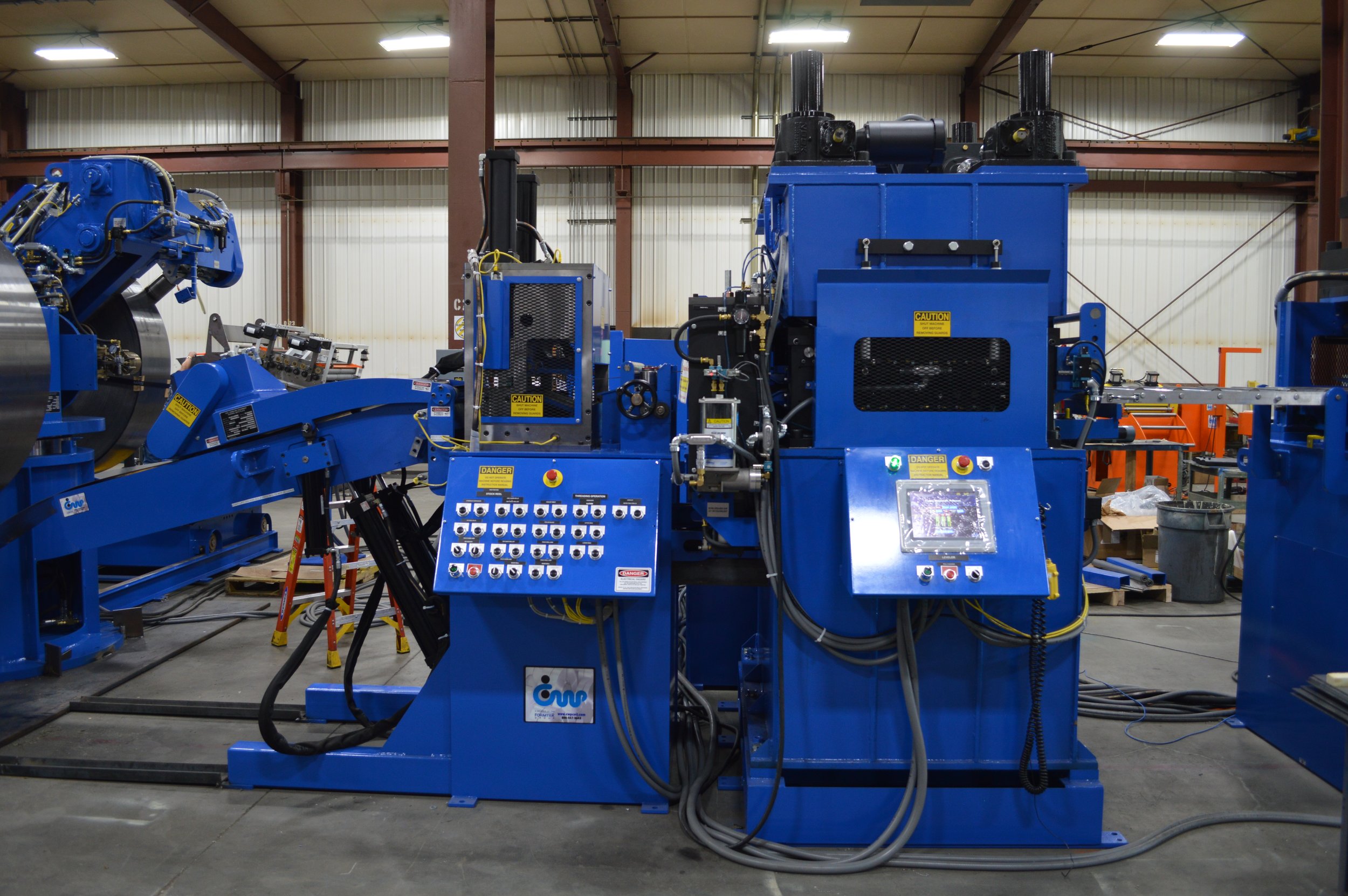 Industrial machinery in a manufacturing workshop with blue equipment, control panel, and various hoses, with metal beams and walls in the background.