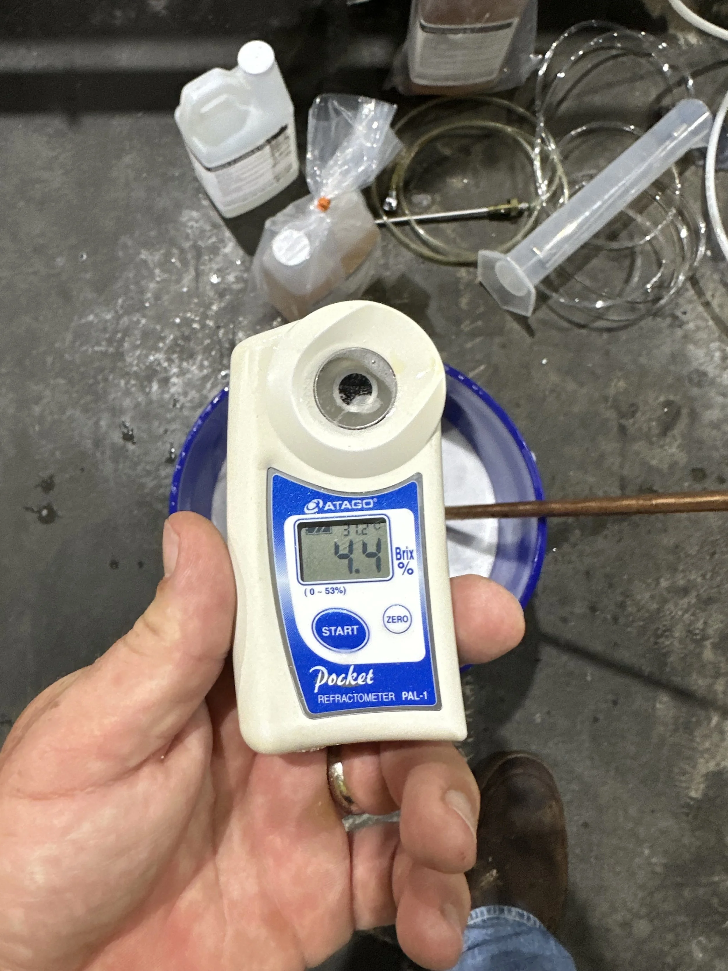 A person is holding a refractometer reading 4.4 Brix, used for measuring sugar content, with laboratory equipment and glassware in the background on a concrete surface.