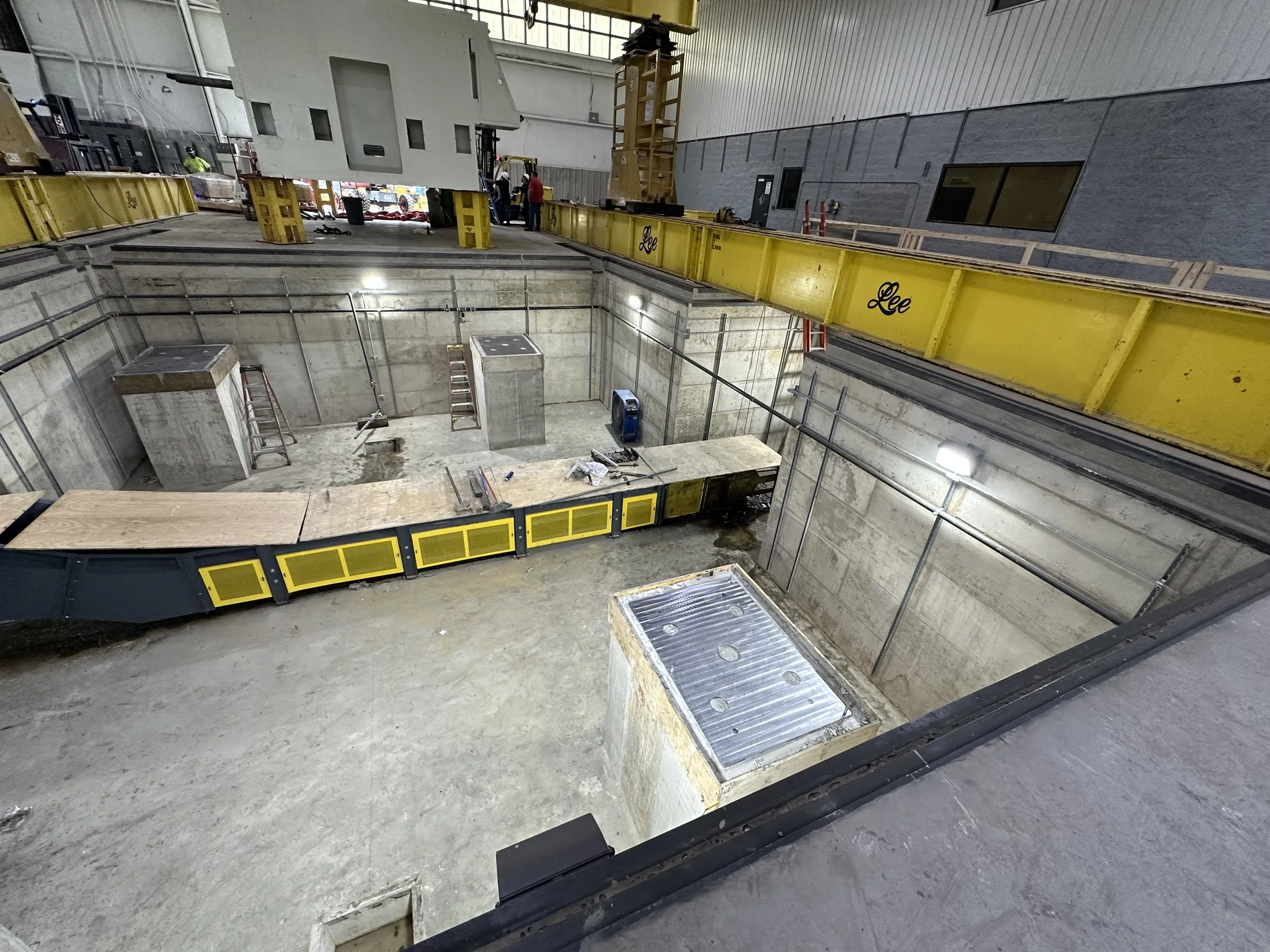 Inside a building under construction with a large rectangular excavation pit. There are yellow safety barriers with the brand name Leo, and various construction tools and equipment are visible around the site.