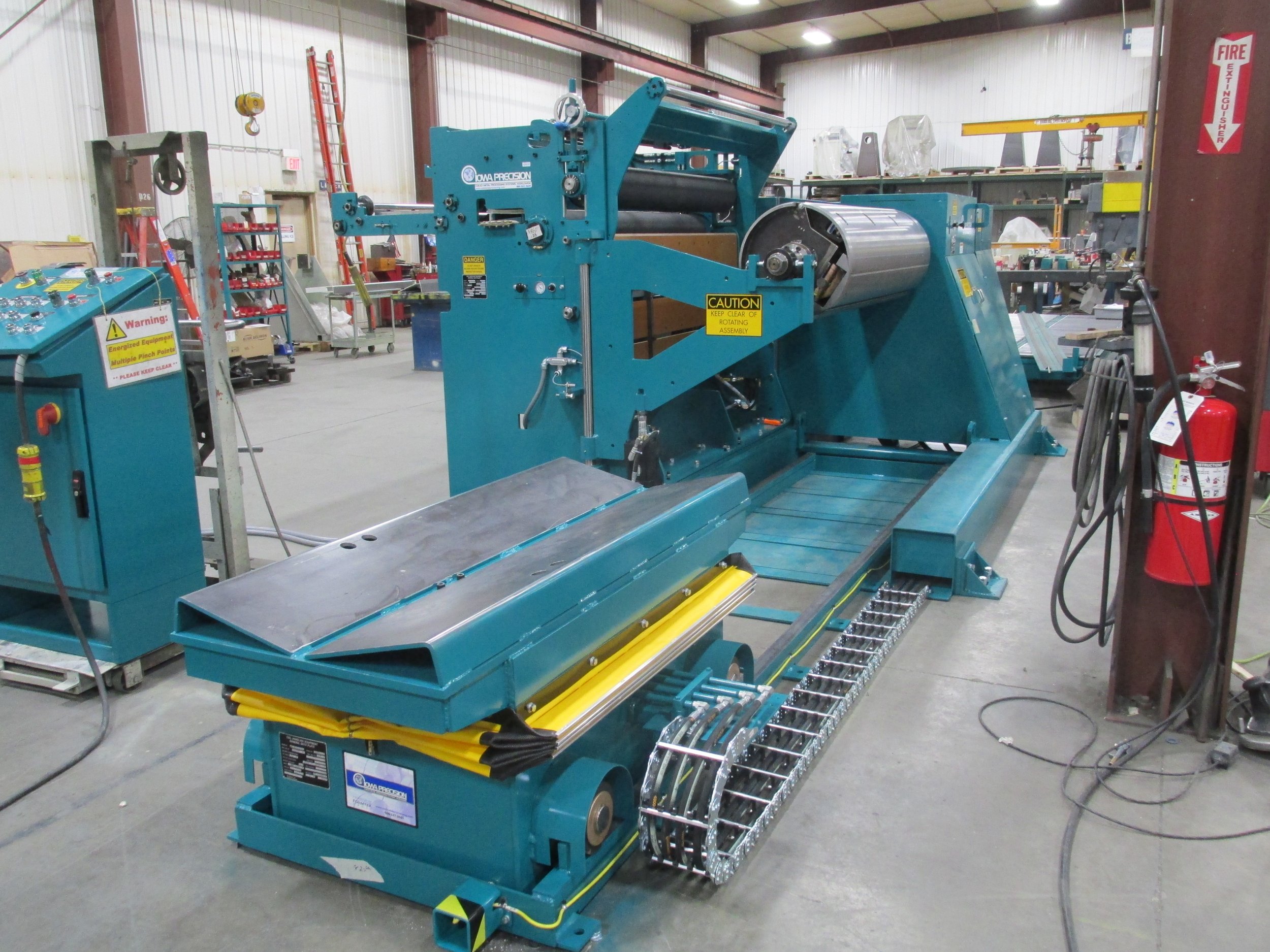 Large industrial machine with conveyor and rollers in a workshop, labeled with caution and warning signs, surrounded by tools and safety equipment.