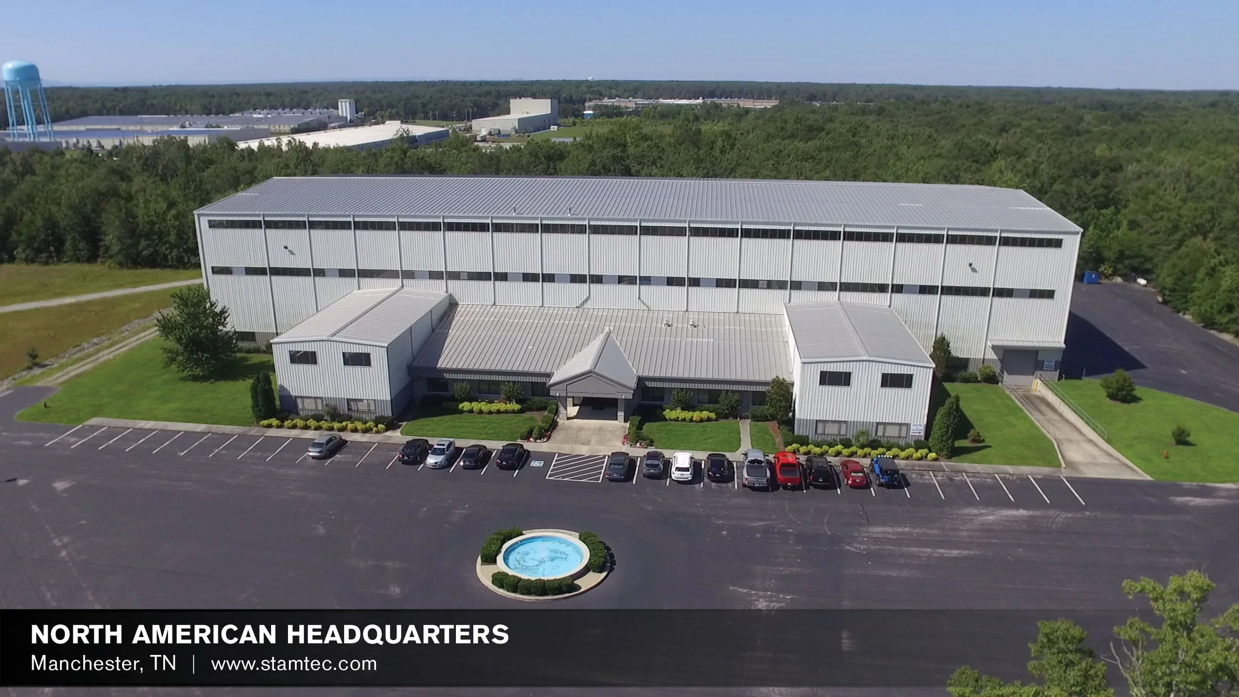 Aerial view of North American Headquarters building in Manchester, TN, showing a large gray industrial-style building, a parking lot with cars, a small landscaped fountain, and surrounding green trees.