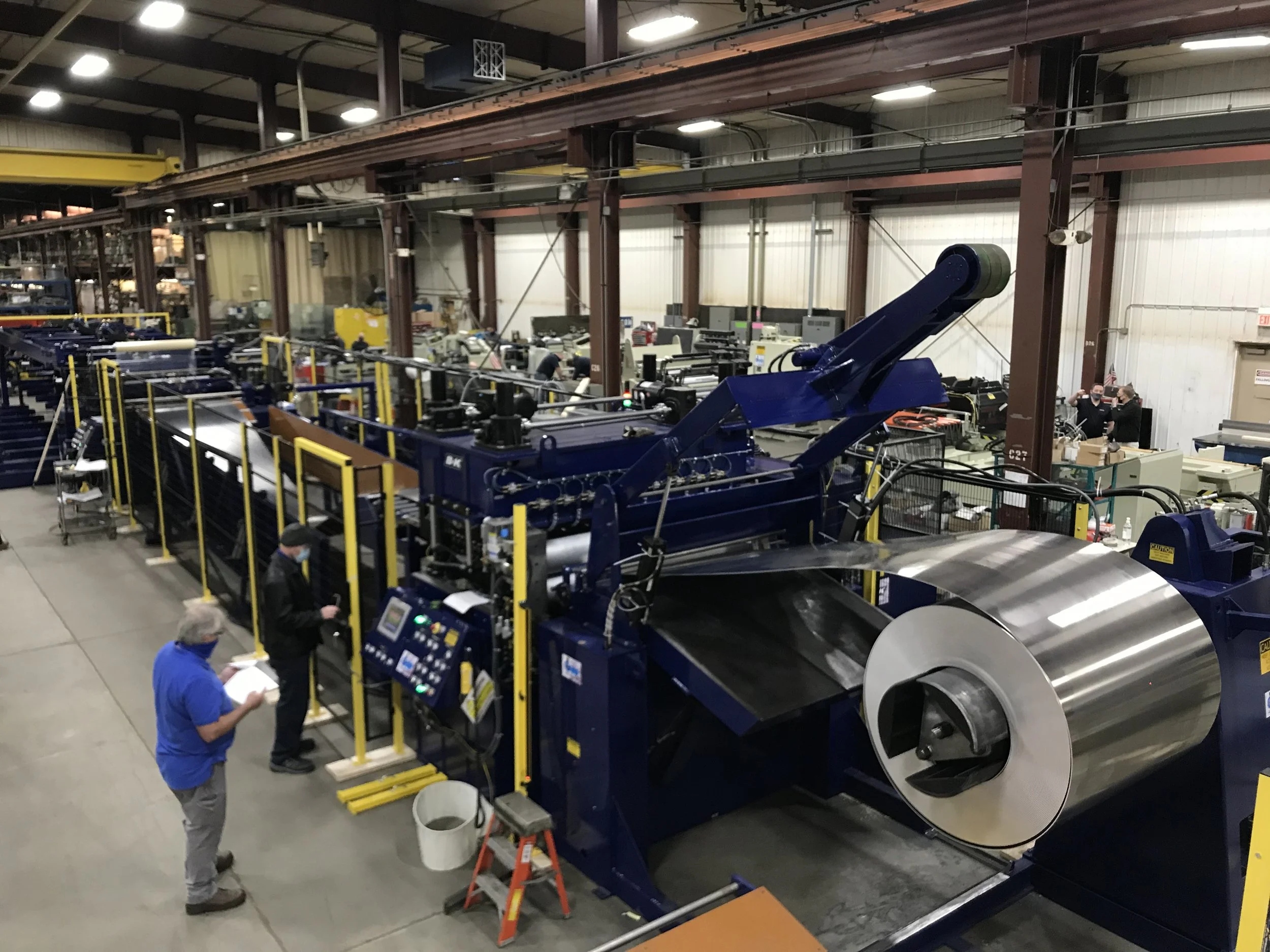 Large industrial manufacturing facility with machinery, including a large roll of shiny metal, control panels, and workers monitoring operations.