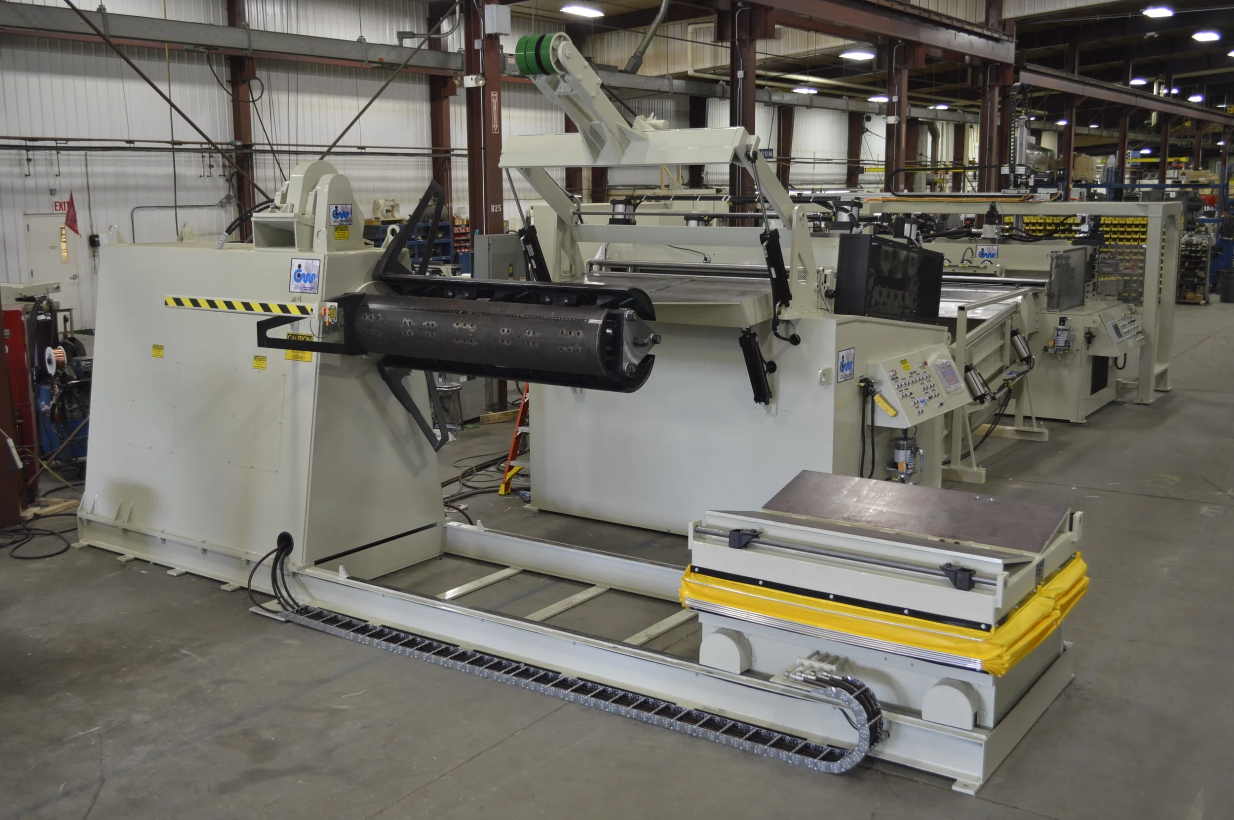 Industrial manufacturing equipment inside a factory, including automated machinery with rollers, control panels, and conveyor systems.
