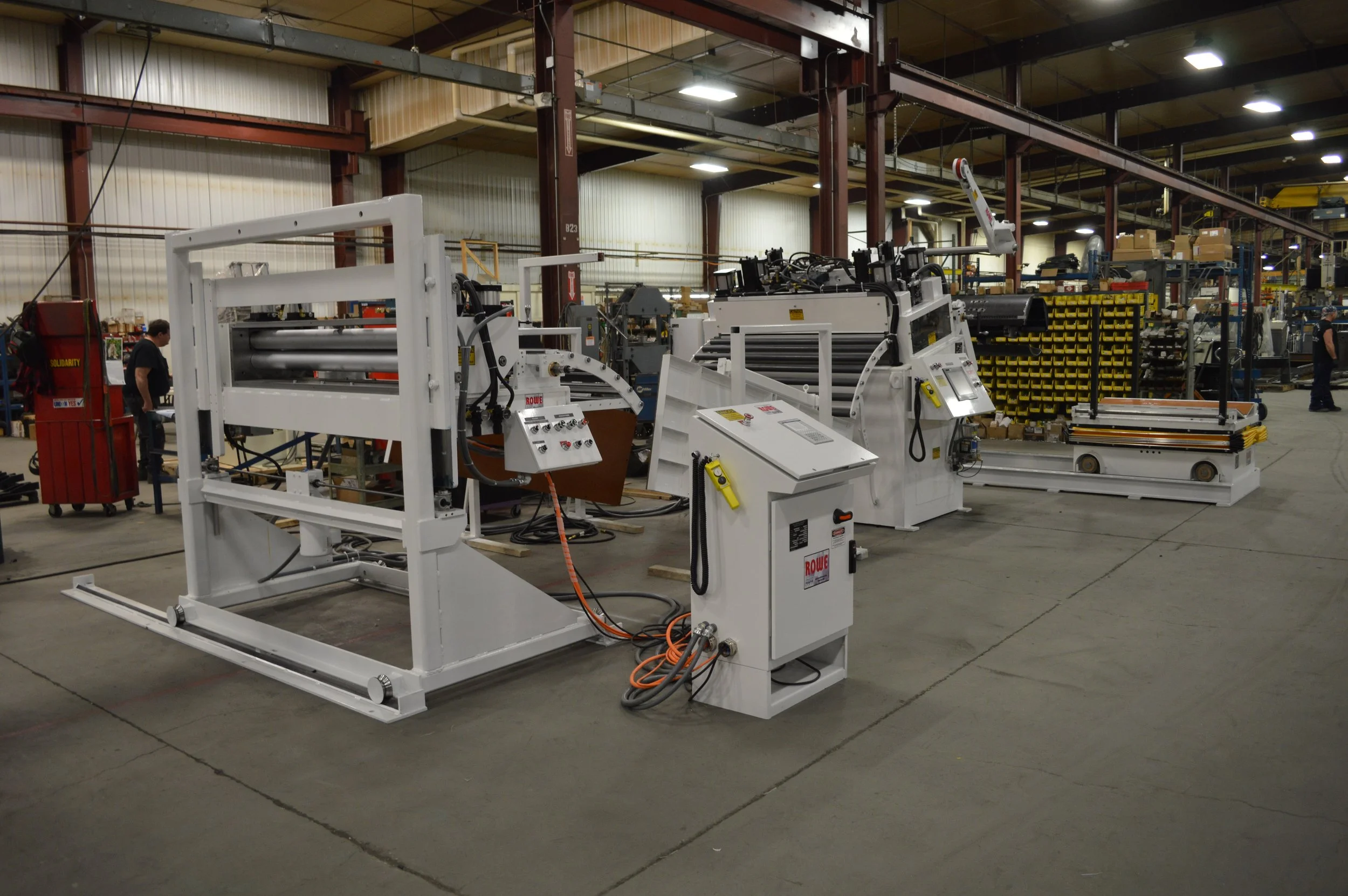 Industrial manufacturing machinery inside a large factory with a concrete floor, metal beams, and shelves of yellow bins in the background.
