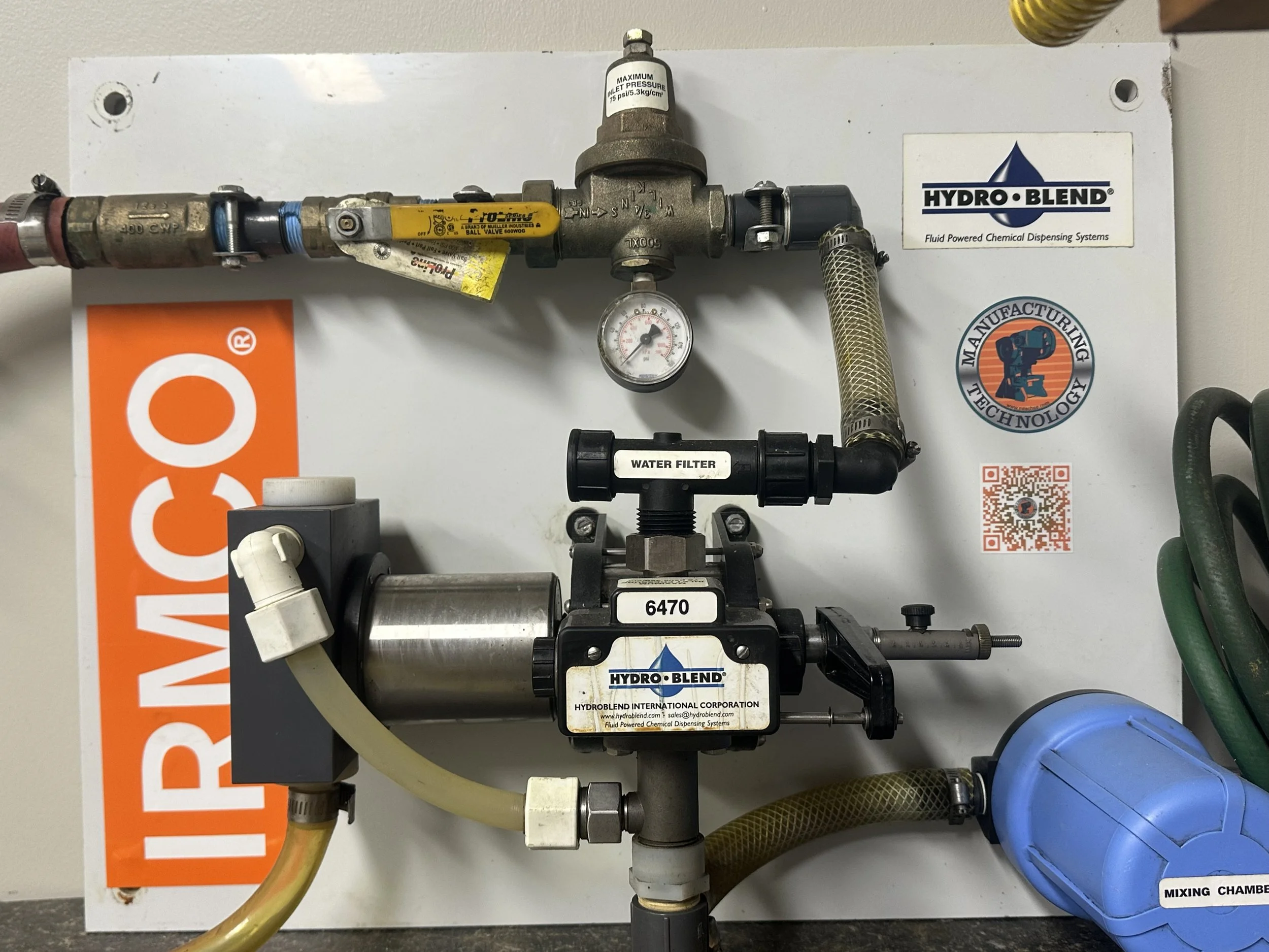 Industrial chemical dispensing system with labeled water filter, mixing chamber, and various pipes and gauges mounted on a white panel.