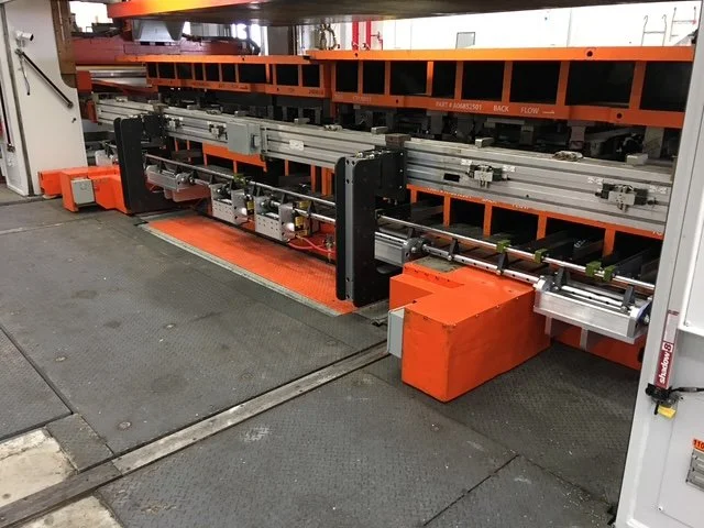 Industrial manufacturing machine with orange and black components, metal rails, and electronic controls situated on a factory floor.