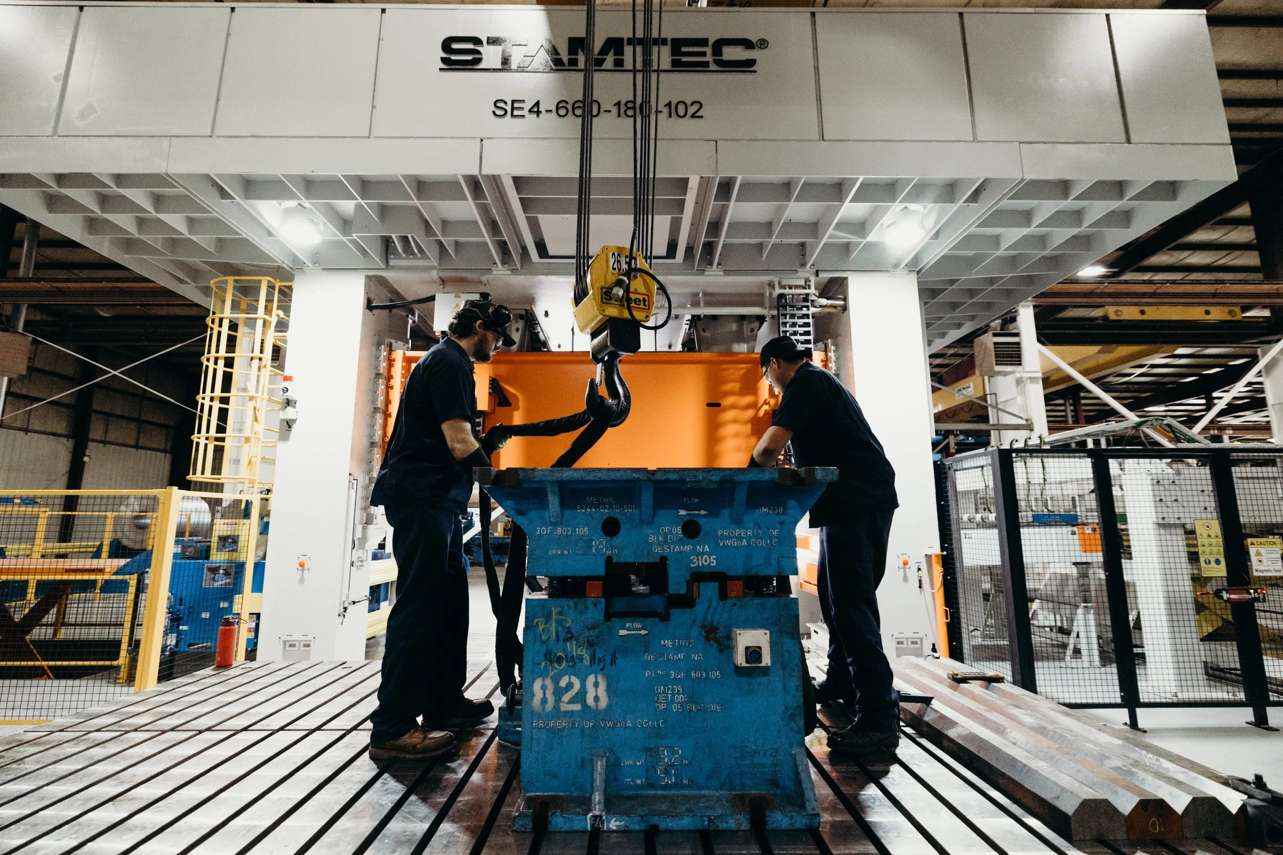 Two workers in dark uniforms and protective gear lifting a large, curved metal piece with a crane in an industrial manufacturing plant. The background shows equipment, safety barriers, and a large overhead structure with the brand name 'STAMTEC'.