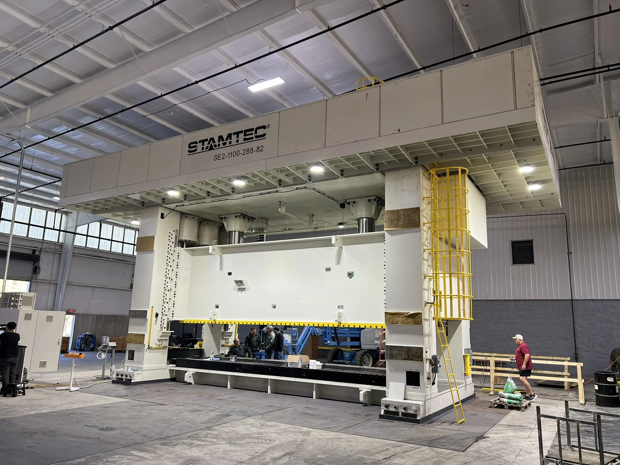 Large industrial machine in a warehouse with several workers around and a man wearing a red shirt and white cap standing on protective mats.