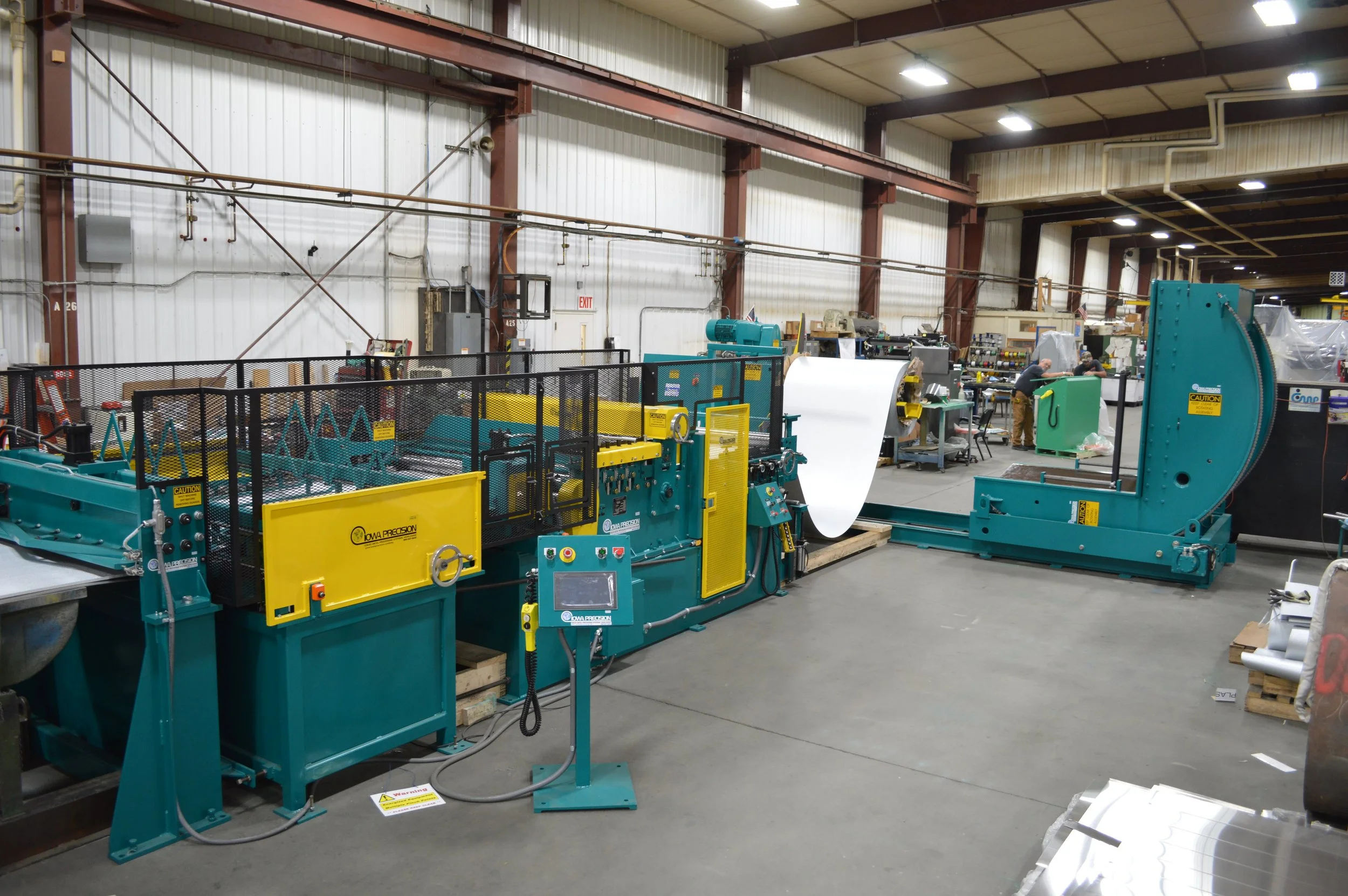 Industrial manufacturing facility with large teal and yellow machinery, including a device feeding a roll of material into a machine, and another large teal machine with a curved arm. Workers in the background operate equipment, and the space has hig