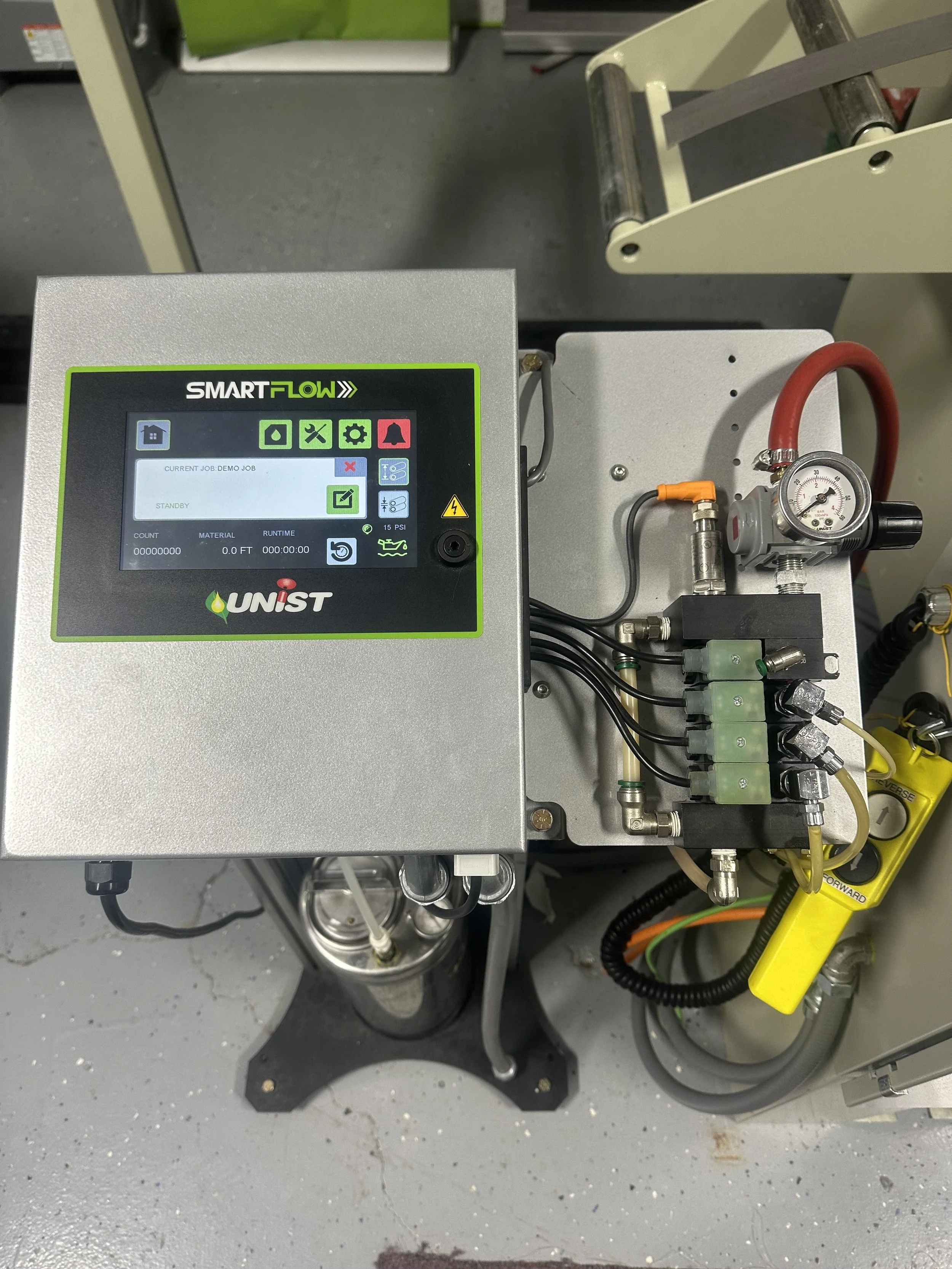 An industrial control panel with a digital screen labeled 'SMARTFLOW' connected to various hoses, valves, and gauges, situated on a gray concrete floor.