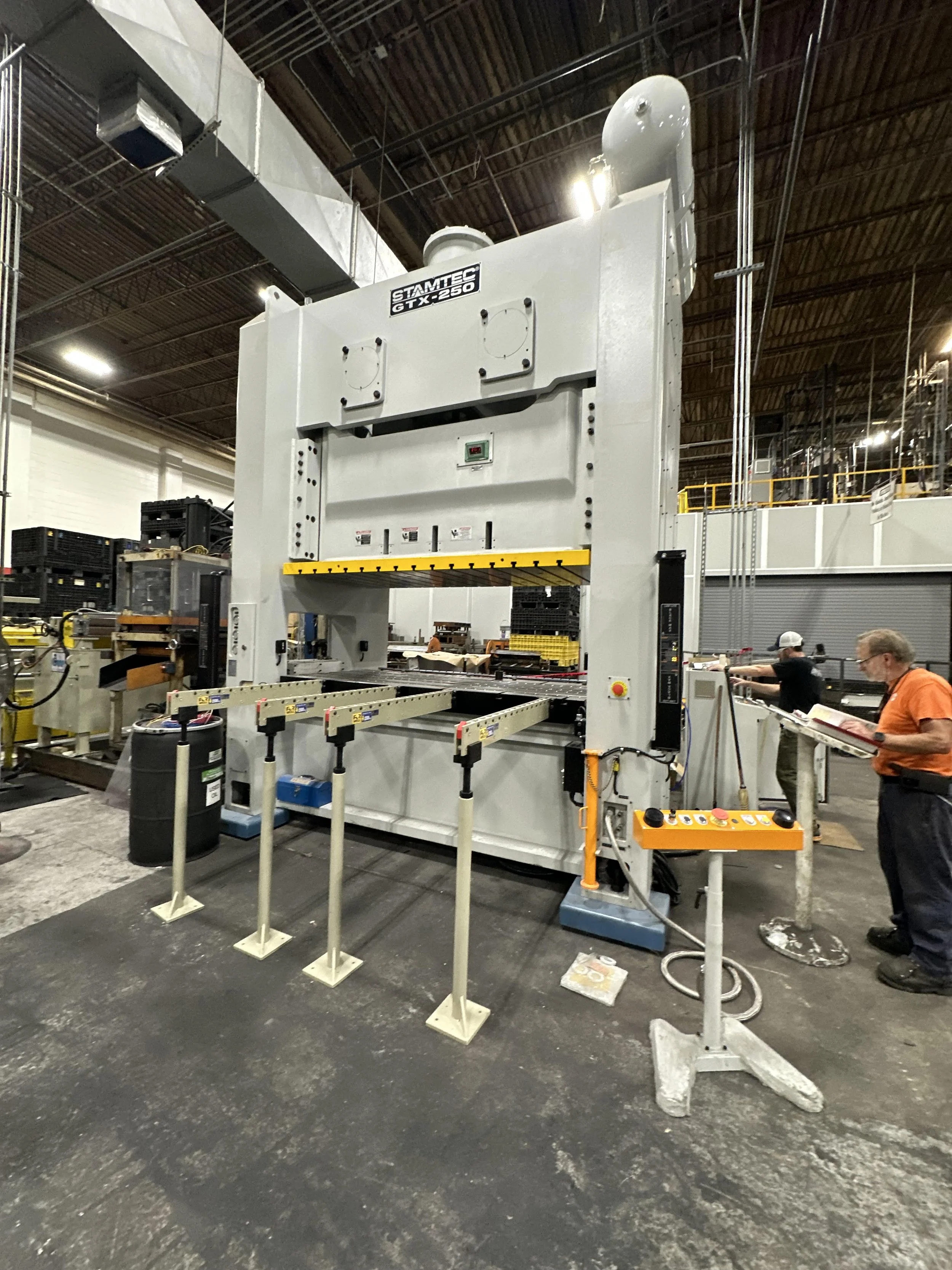 Large industrial machine in a factory setting labeled 'STAMTEC GTX 250'. Several workers are operating the machine and inspecting it.