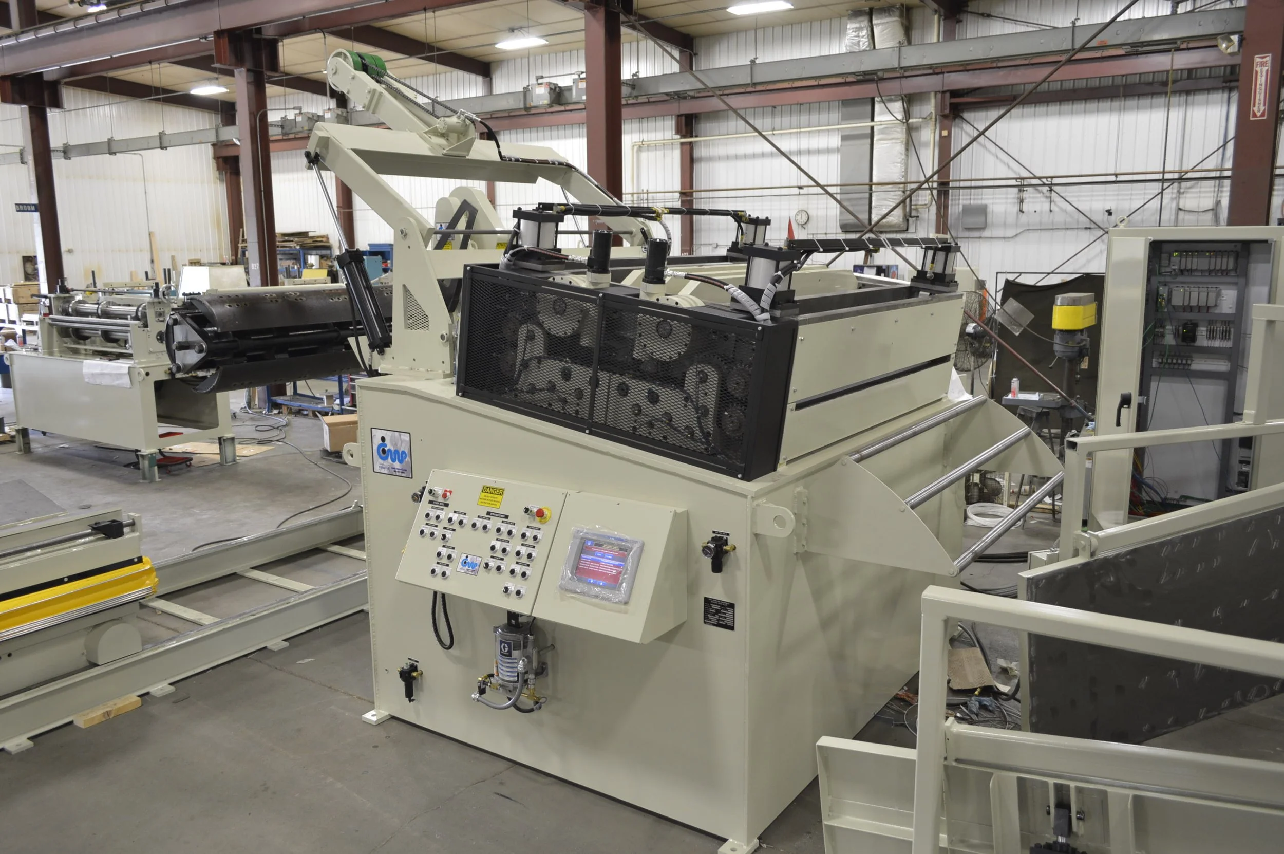 Industrial machinery in a factory setting, including a large white machine with control panels and a black mesh section, surrounded by other equipment and metal framework.
