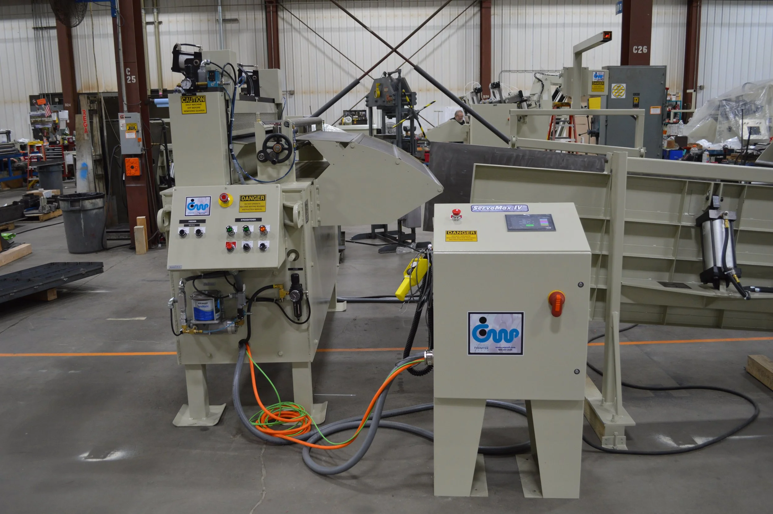 Industrial machine setup in a factory with control panels, wiring, and safety signs, with a person working in the background.