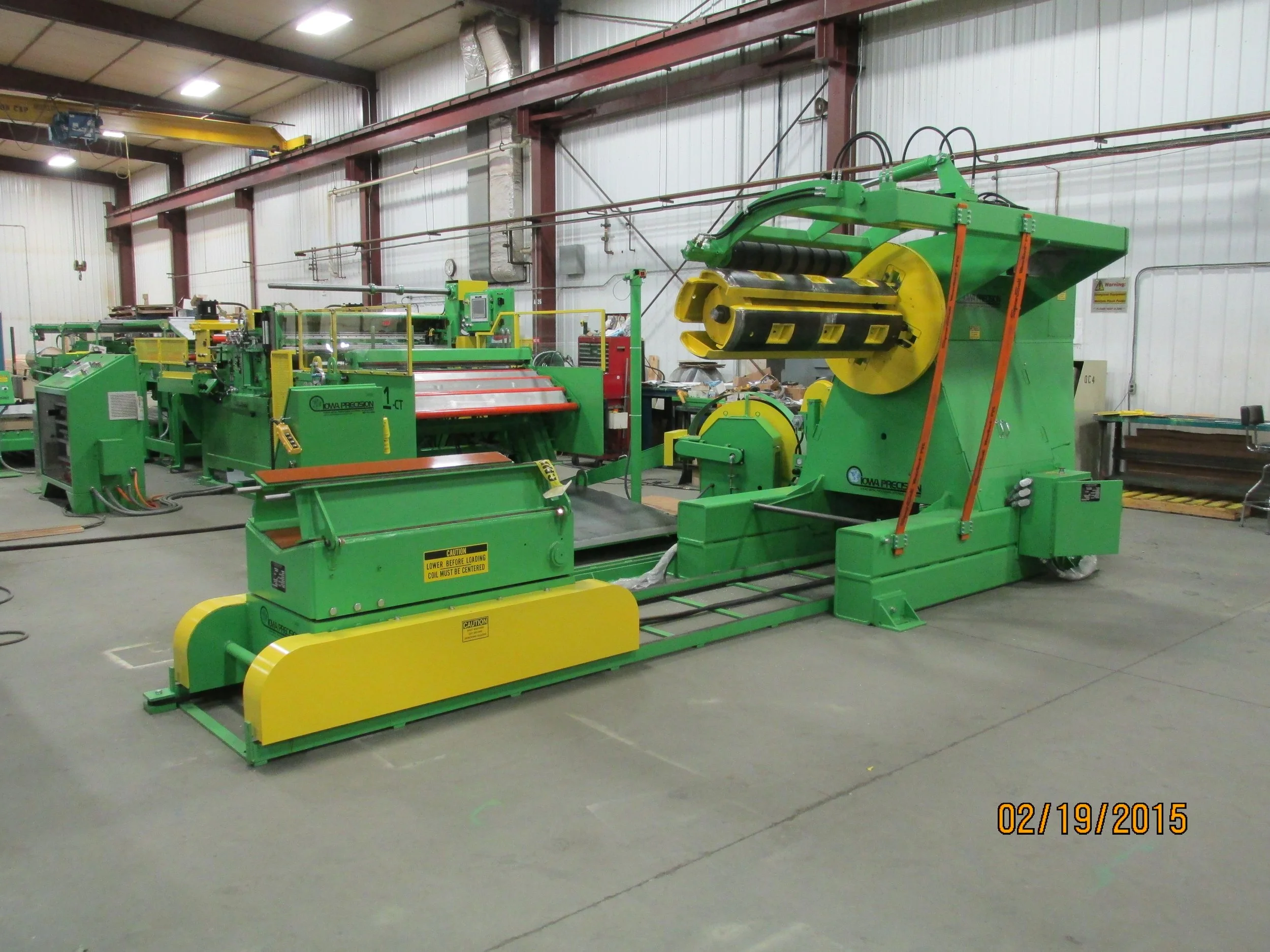 Industrial machinery in a factory setting, primarily green and yellow, with various rollers, panels, and control systems, used for manufacturing or processing materials.