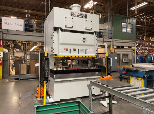 Industrial stamping press machine labeled STAMTEC in a factory setting, with safety barriers and a worktable nearby.