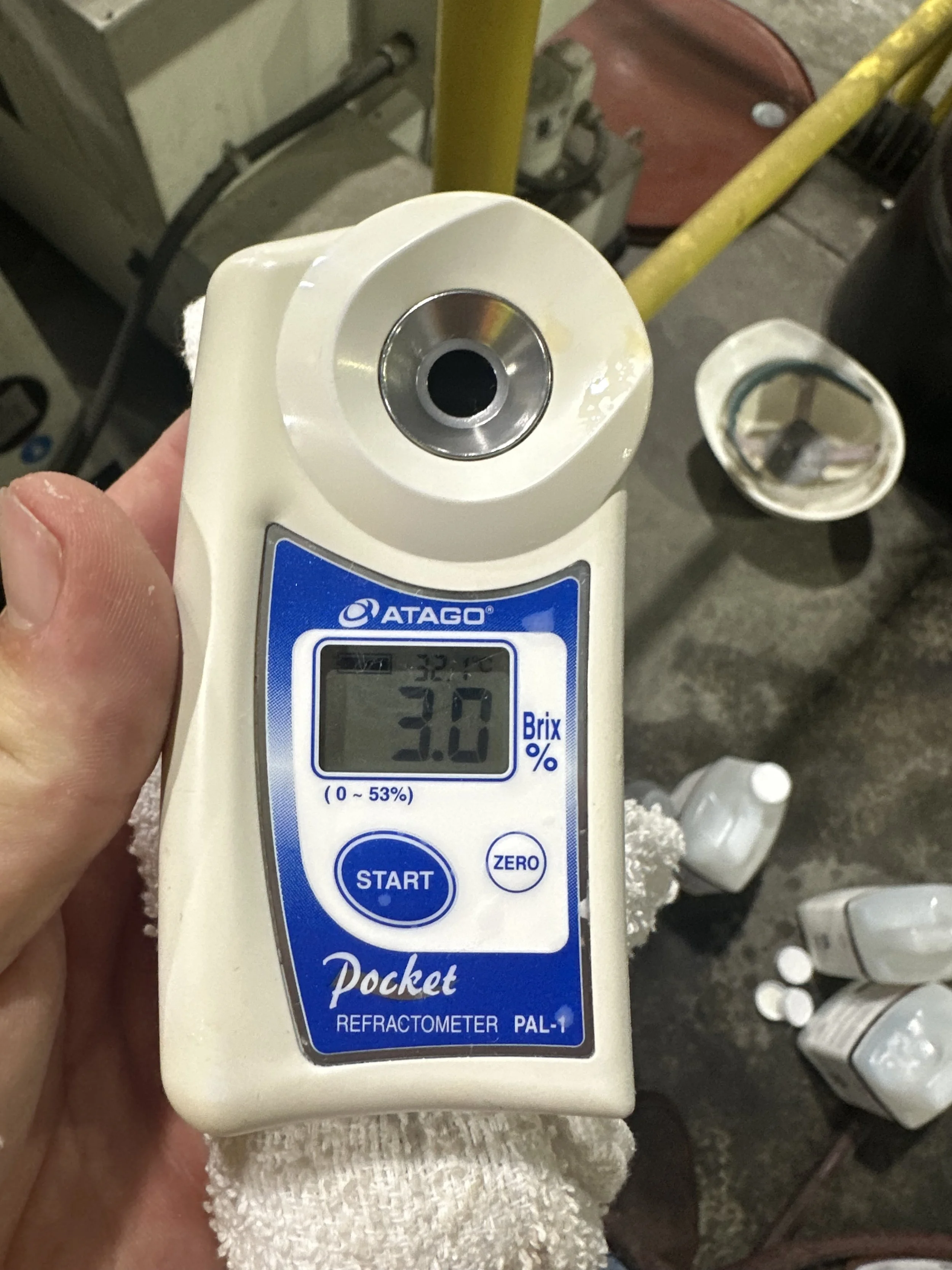 A handheld refractometer showing a Brix percentage of 3.0%. The device is labeled 'Pocket Refractometer PAL-1' and is held by a person wearing a white cloth.