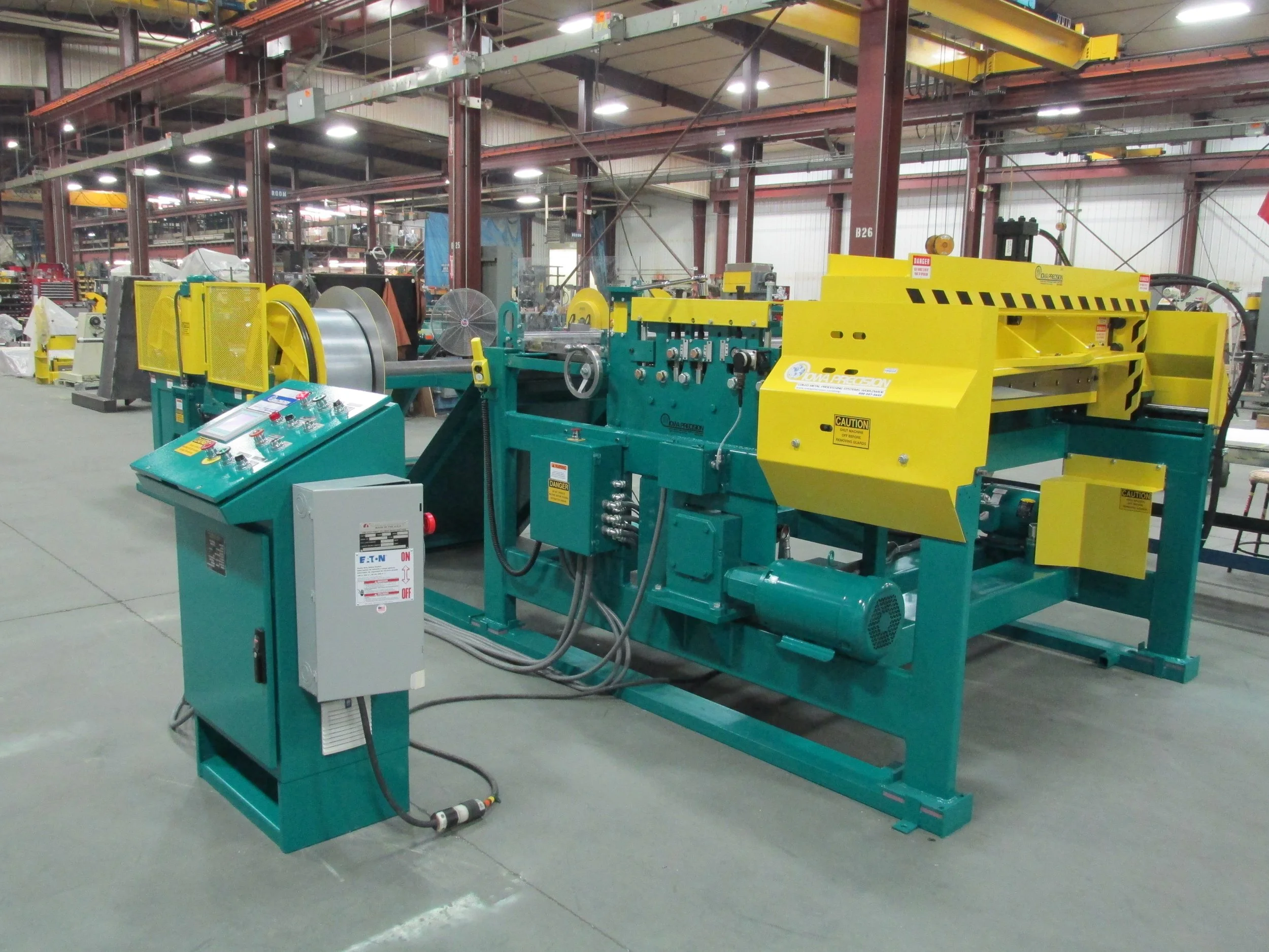 Industrial machine in a factory setting with control panel and mechanical components, painted in teal and yellow.