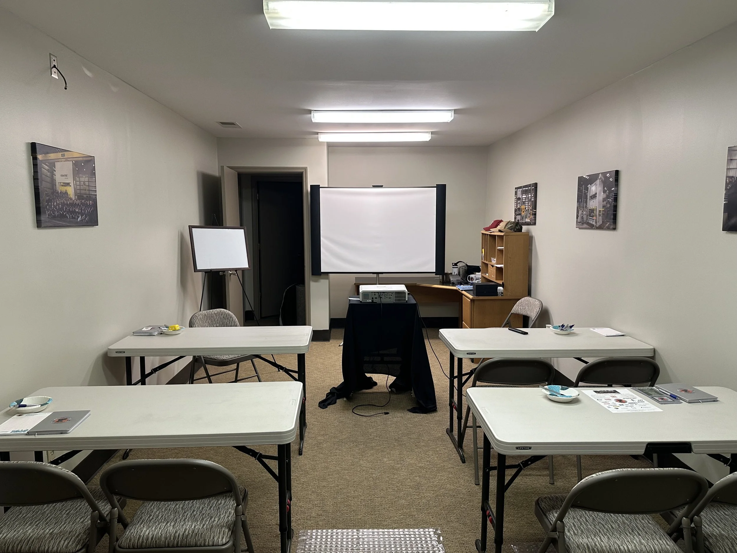 Empty classroom with white desks, folding chairs, a projector, and a projection screen in the front of the room. The walls have framed pictures, and there's a small bookshelf with various items on it.