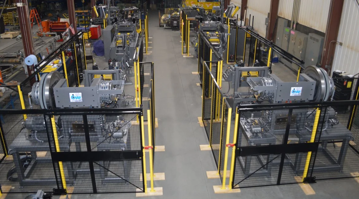 Industrial factory with robotic machines enclosed in yellow and black safety cages, organized in two parallel rows inside a warehouse.