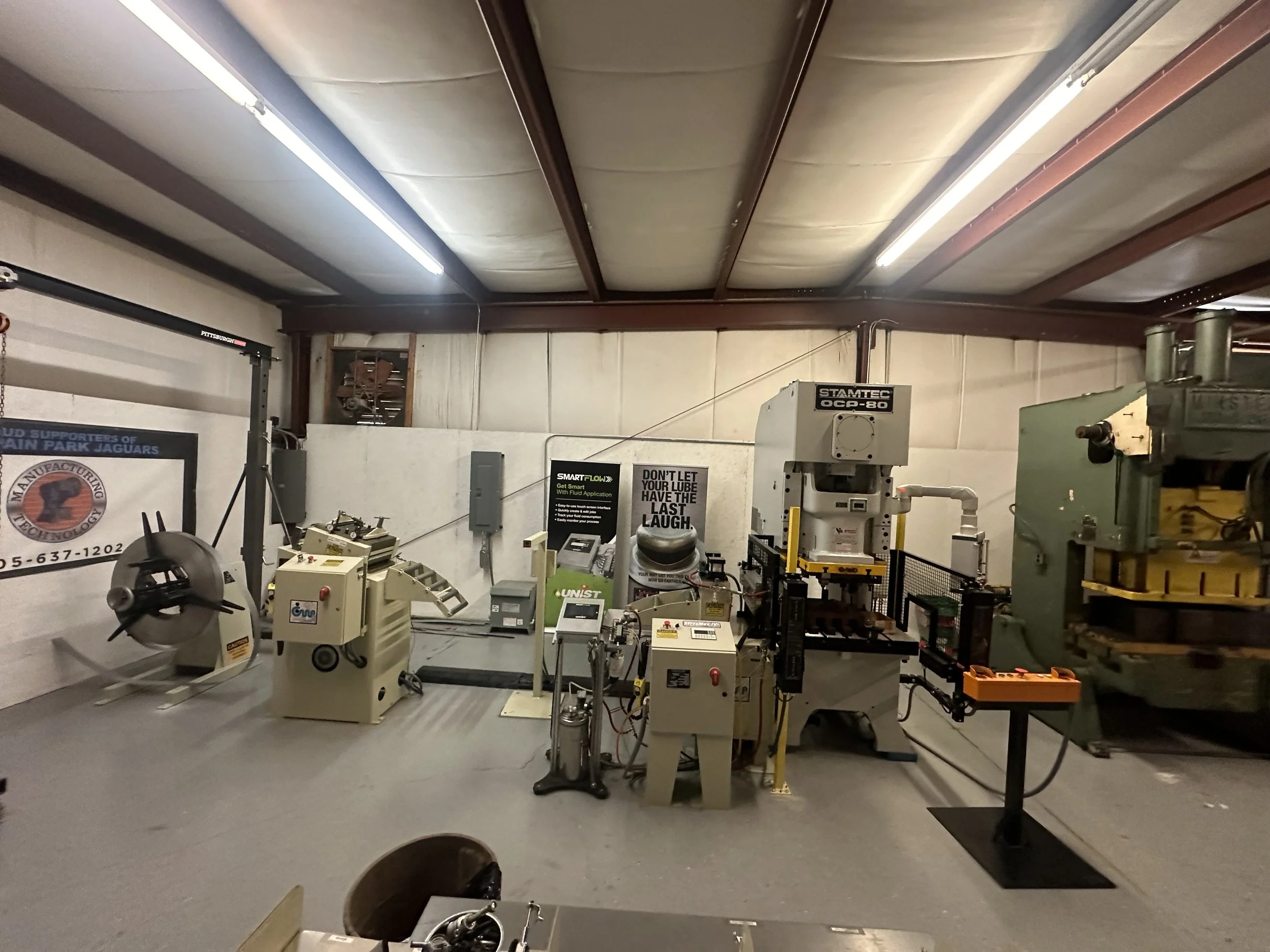 Industrial workshop with machinery, including a metal shear, hydraulic press, and testing equipment, inside a garage or warehouse space.